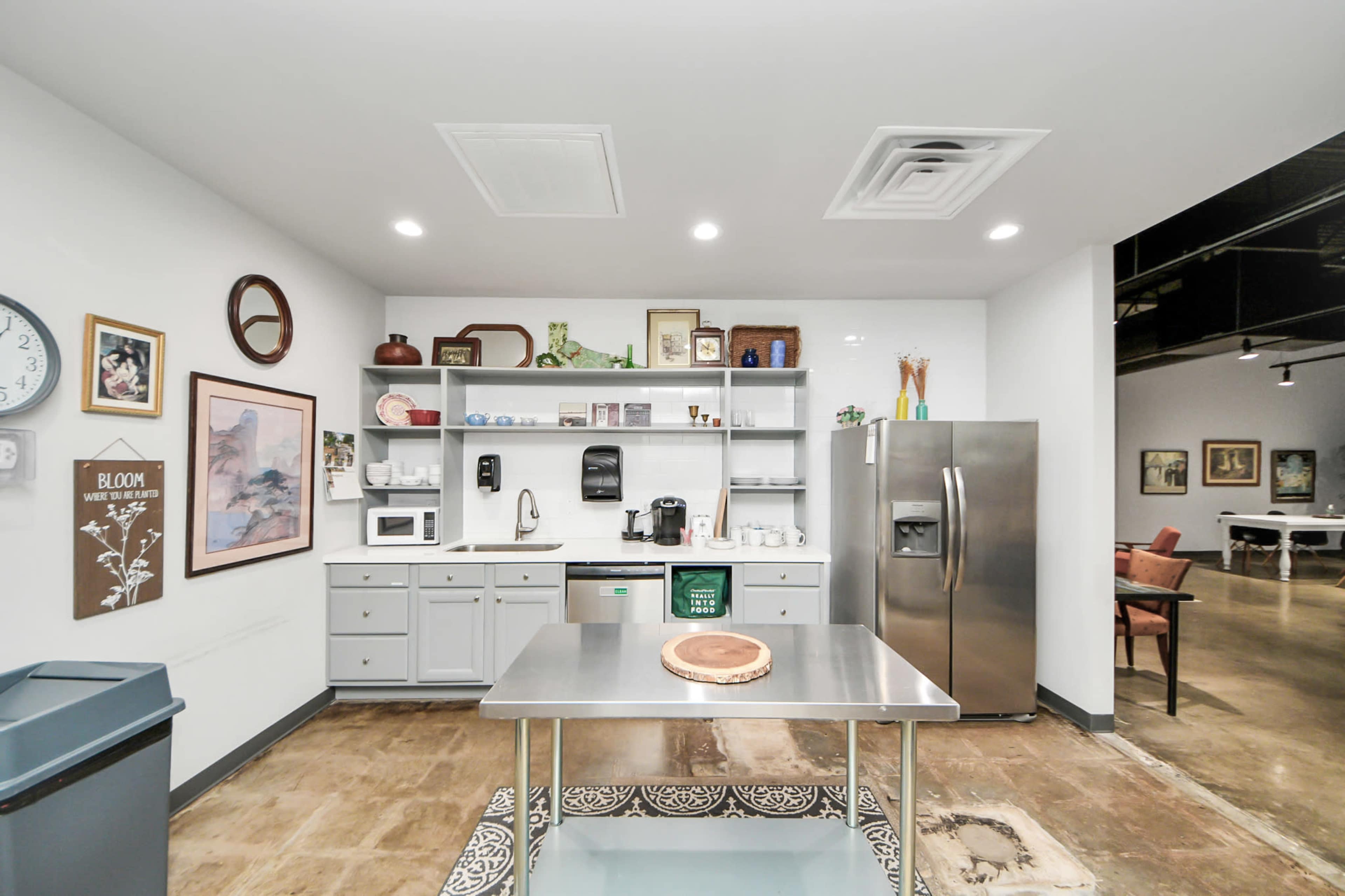 The image shows a modern kitchen setup with gray cabinetry, a stainless steel refrigerator, a stainless steel island, and various kitchen appliances on display against a white wall adorned with framed art.