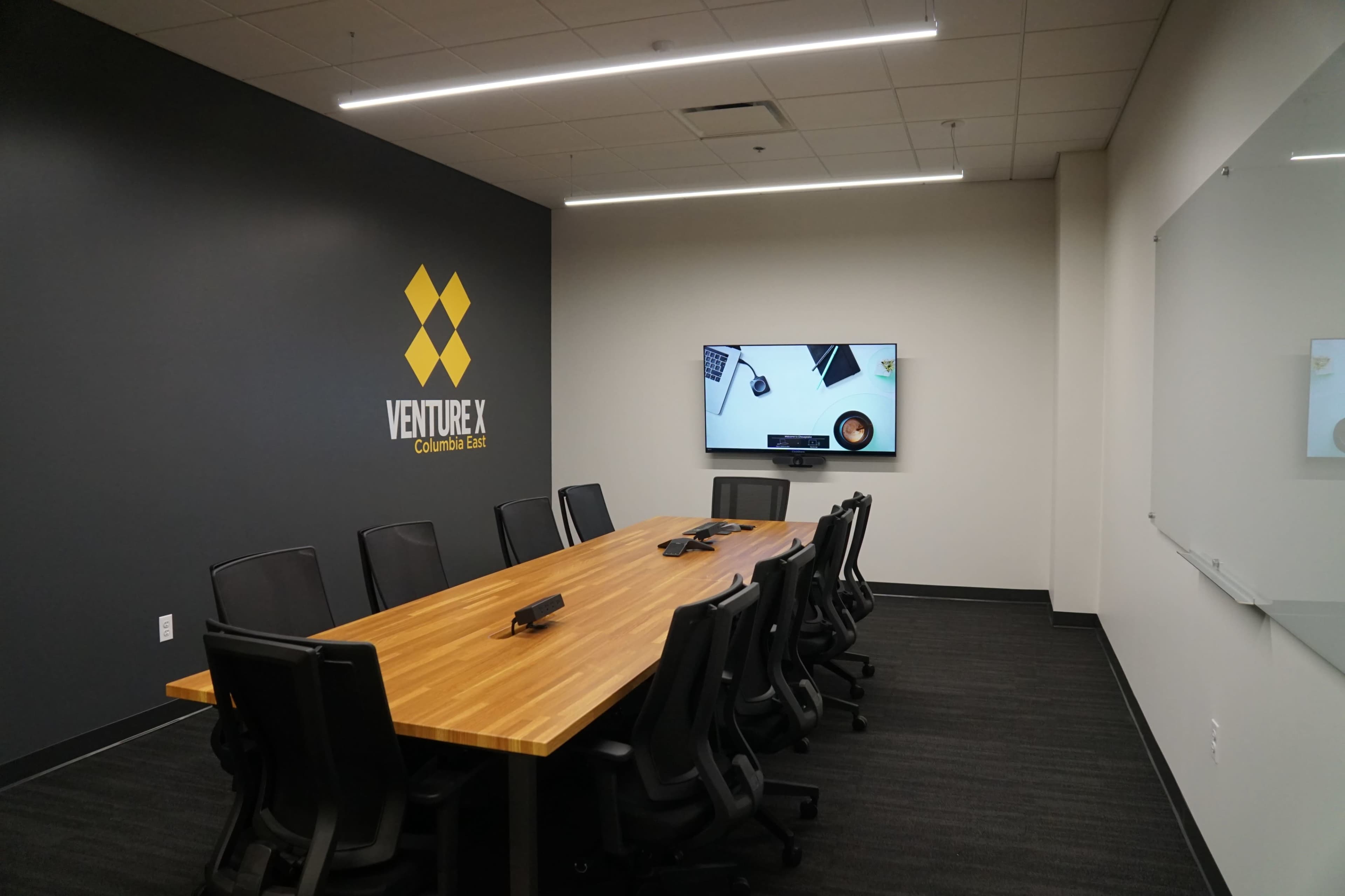 A modern conference room features a large wooden table surrounded by black chairs, a television mounted on the wall, and the "Venture X" logo displayed prominently.
