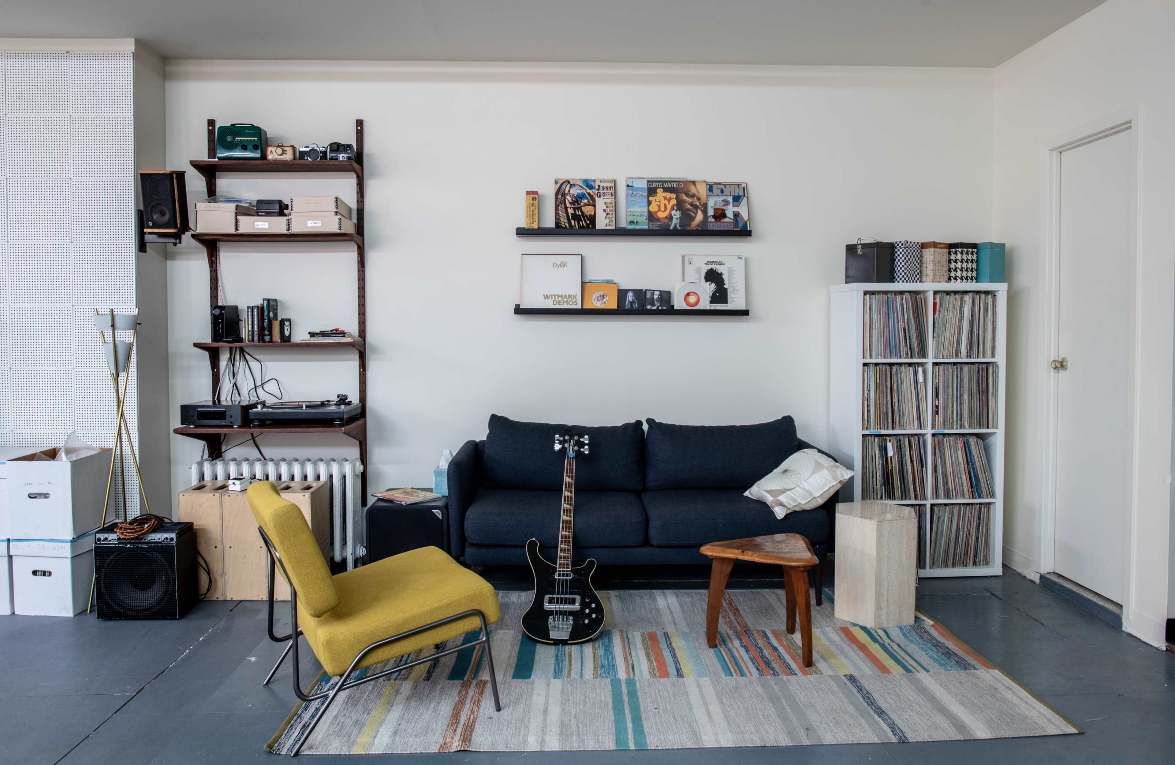 The image shows a living room featuring a blue sofa, a yellow chair, a wooden side table, a record player, vinyl records, and a shelf displaying various items.