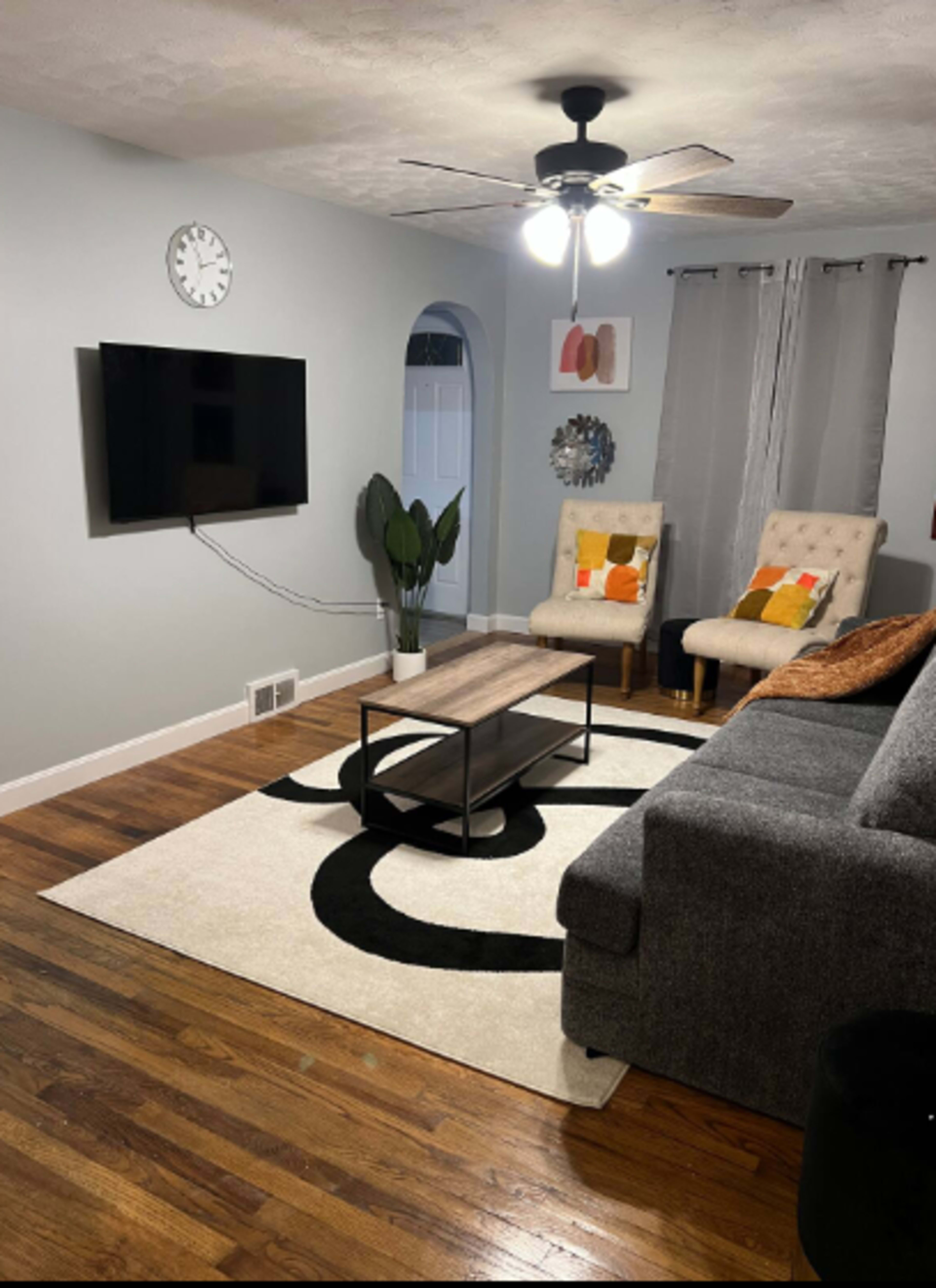 The living room features a gray sofa, two armchairs with colorful cushions, a coffee table, a large TV mounted on the wall, and a tall plant beside a window.