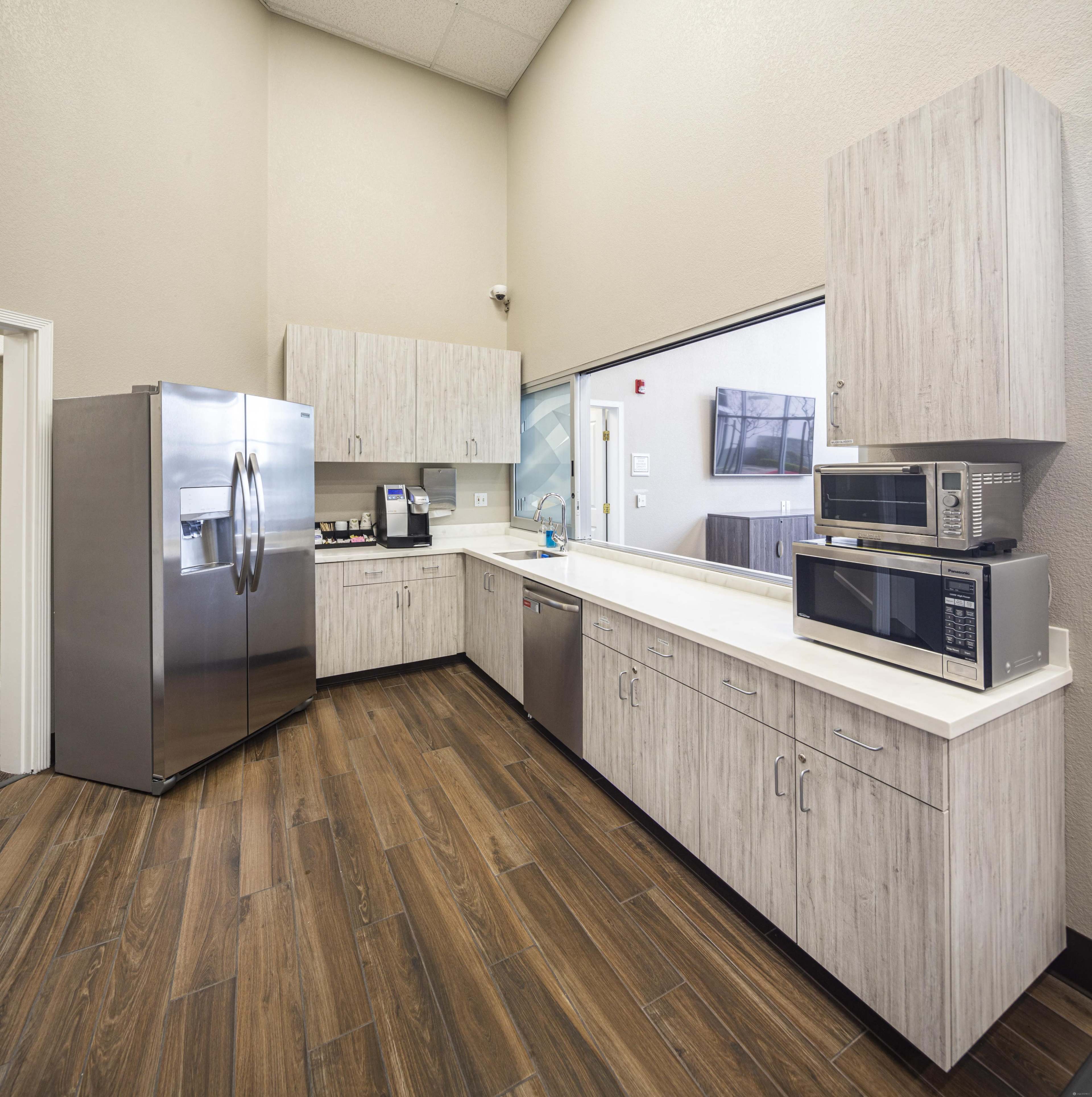 A modern kitchen with stainless steel appliances, including a refrigerator, microwave, and dishwasher, set against light wood cabinetry and a long countertop.