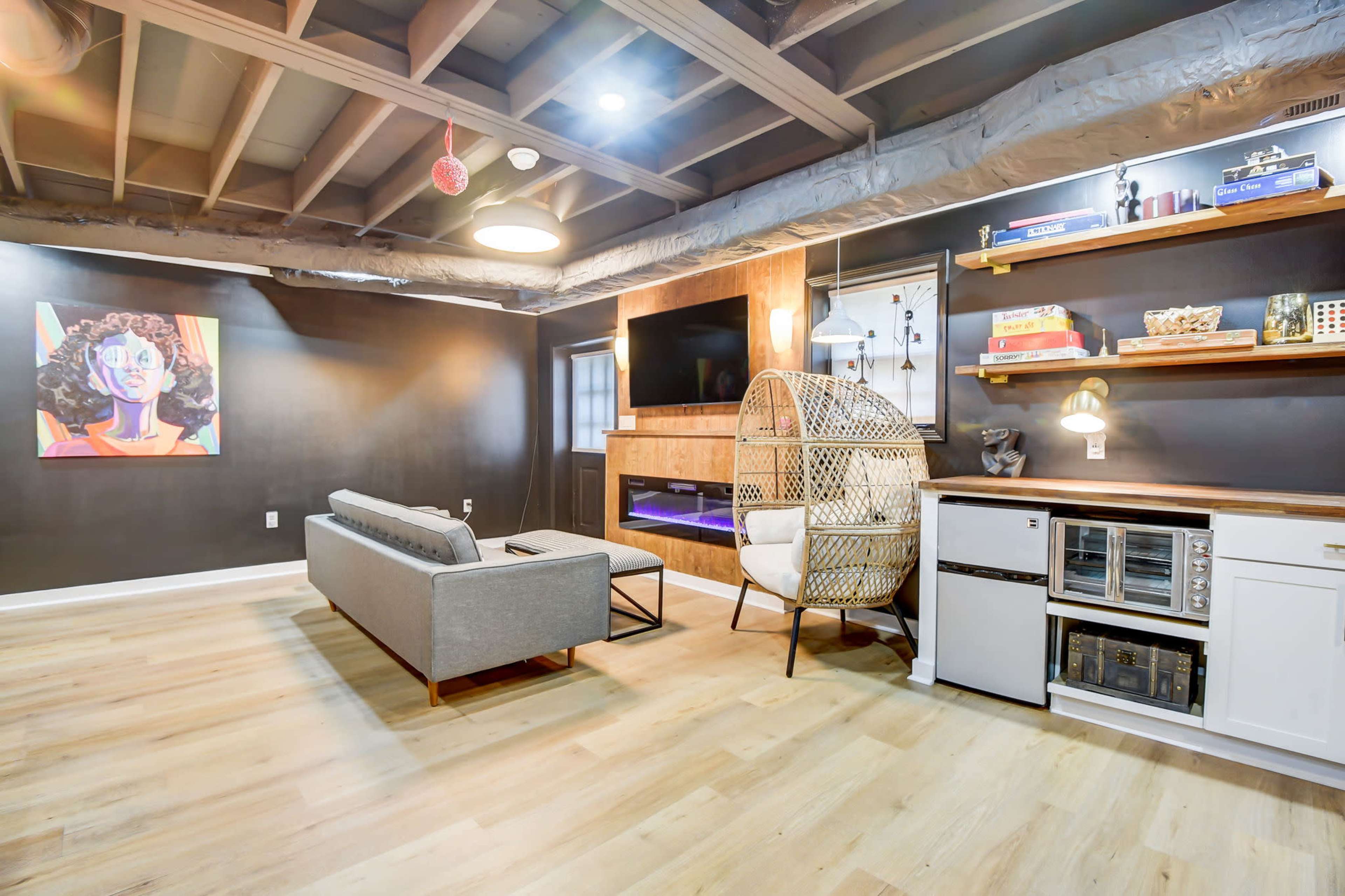 The room features a modern, minimalist lounge area with a gray sofa, a wall-mounted television, and decorative shelves, alongside a cozy chair and a small kitchenette.