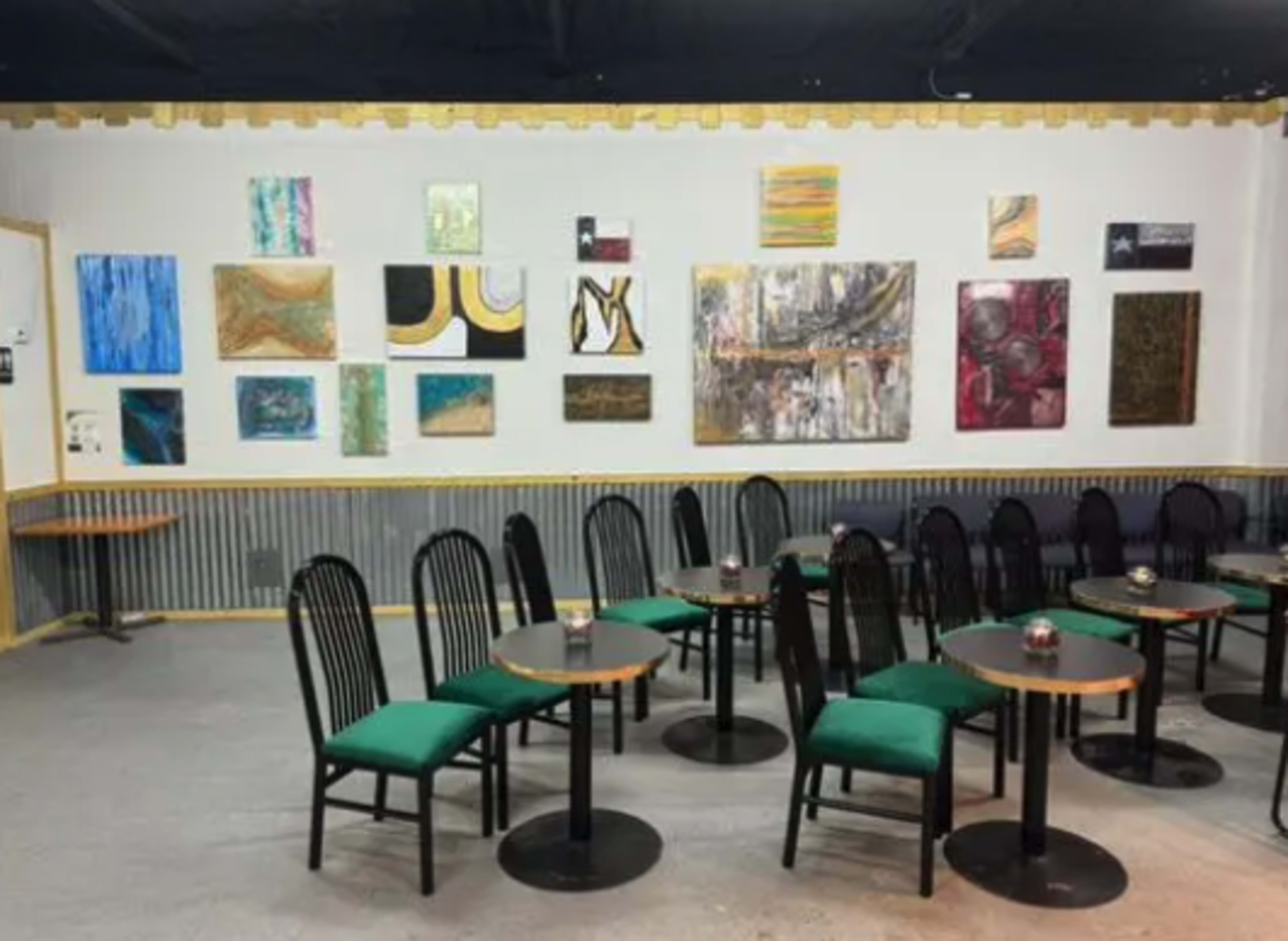 A cafe interior with several small tables and chairs upholstered in green, against a wall decorated with various colorful artworks.
