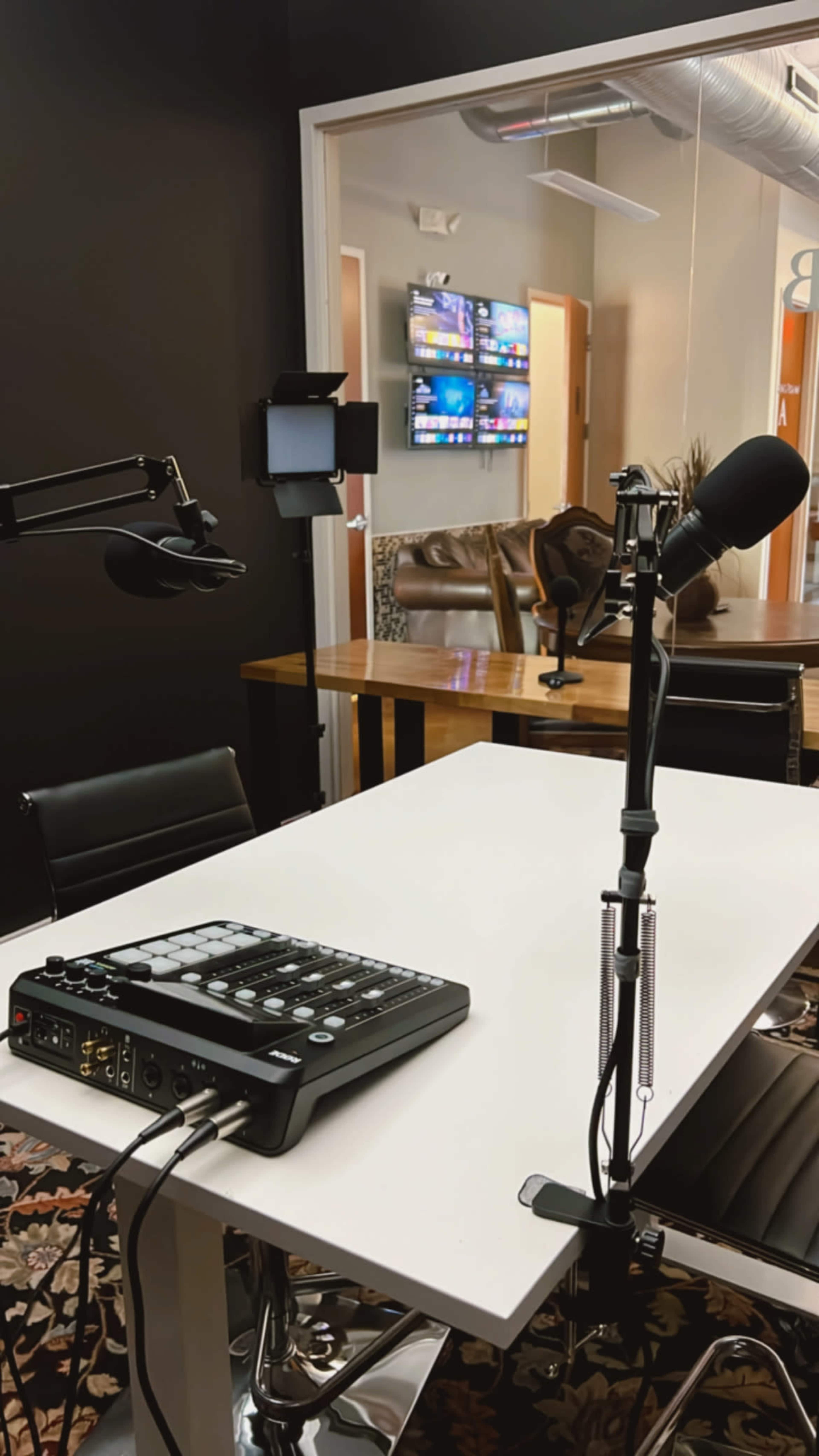 A recording studio features a white table with audio equipment and microphones, set against a dark wall, with a view of a lounge area in the background.