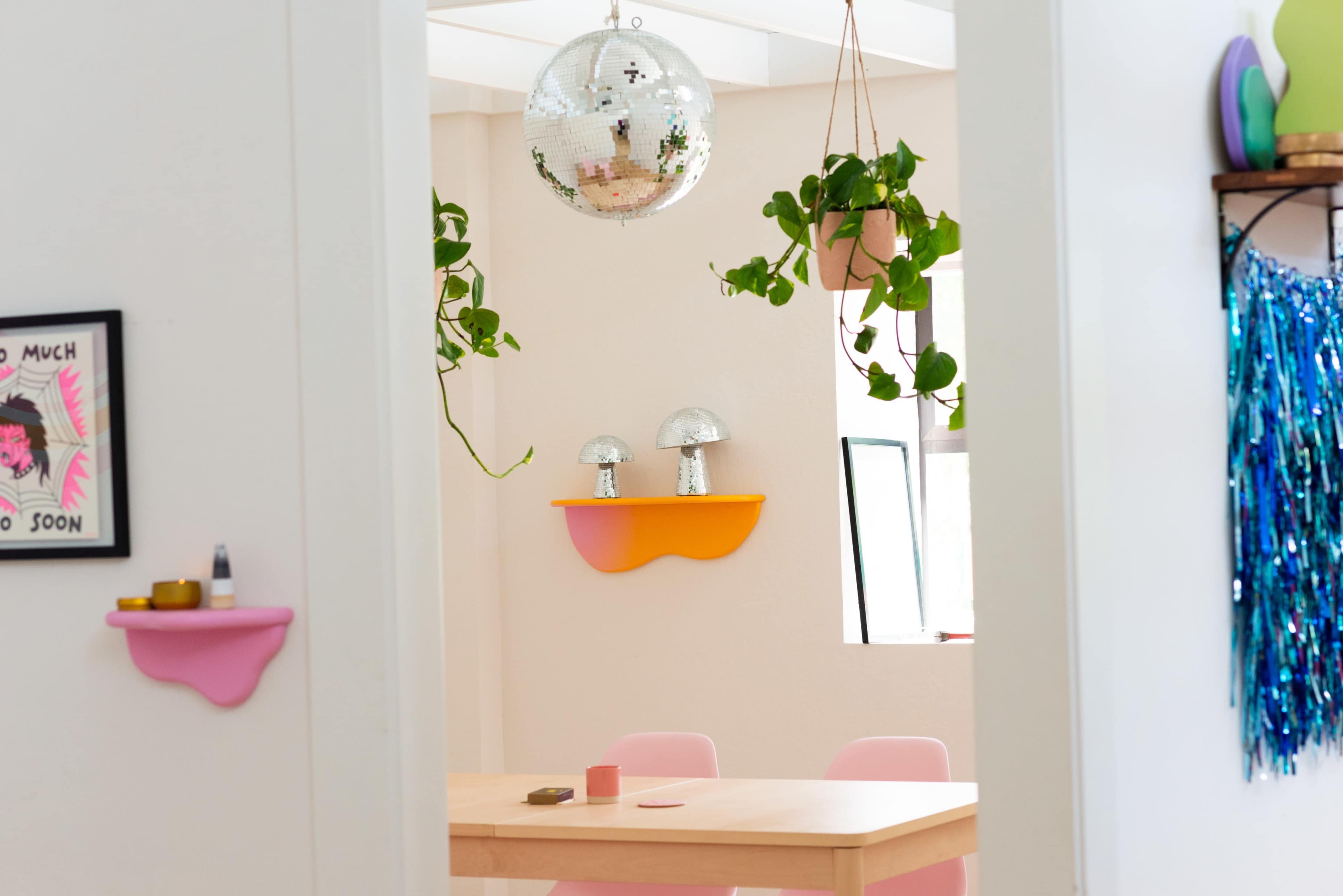 The image shows a bright dining area with a wooden table, pink chairs, and a disco ball hanging from the ceiling, alongside potted plants on shelves.