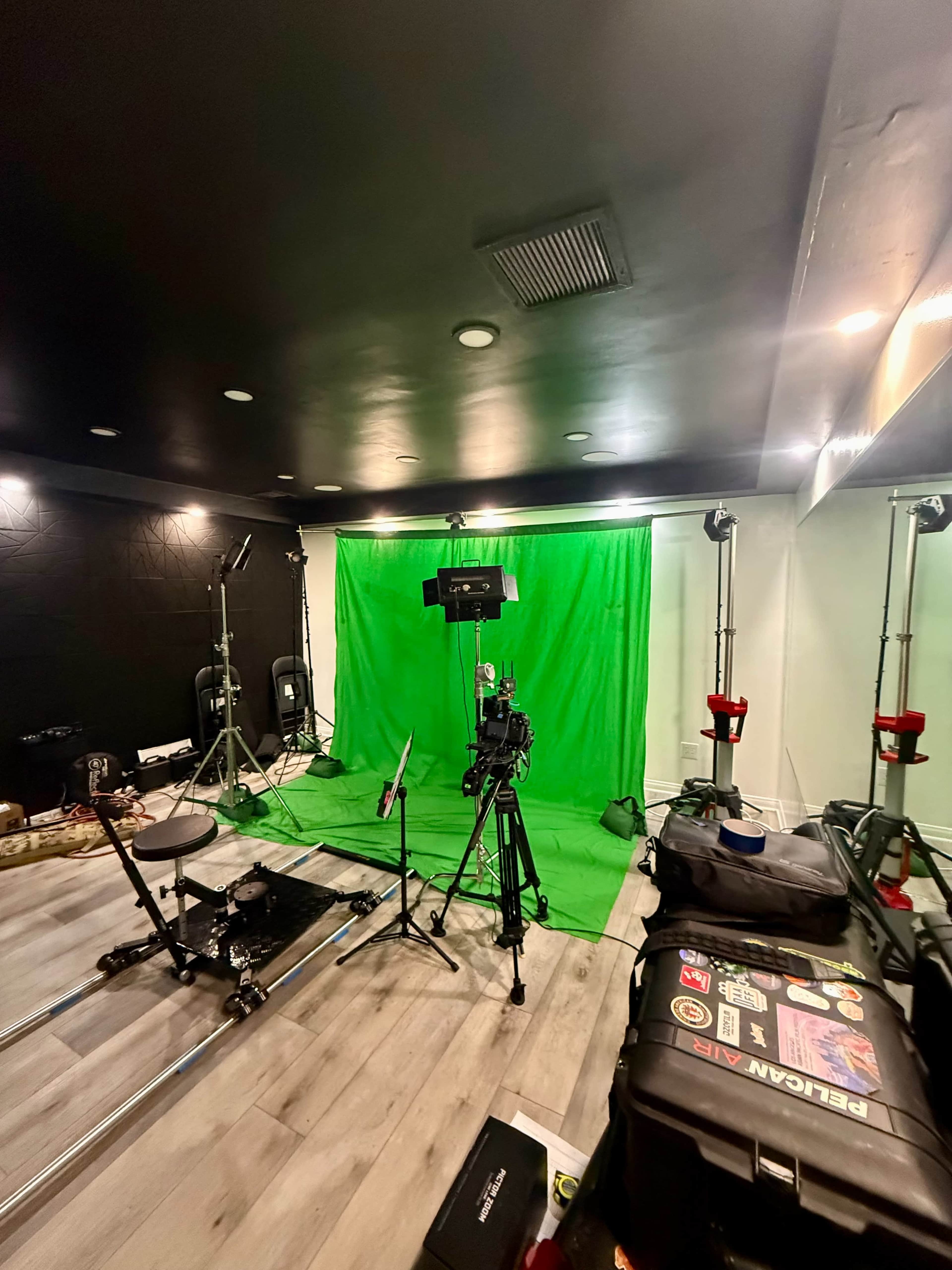 The image shows a studio setup featuring a green screen, cameras on tripods, and lighting equipment in a room with a dark ceiling and wooden flooring.