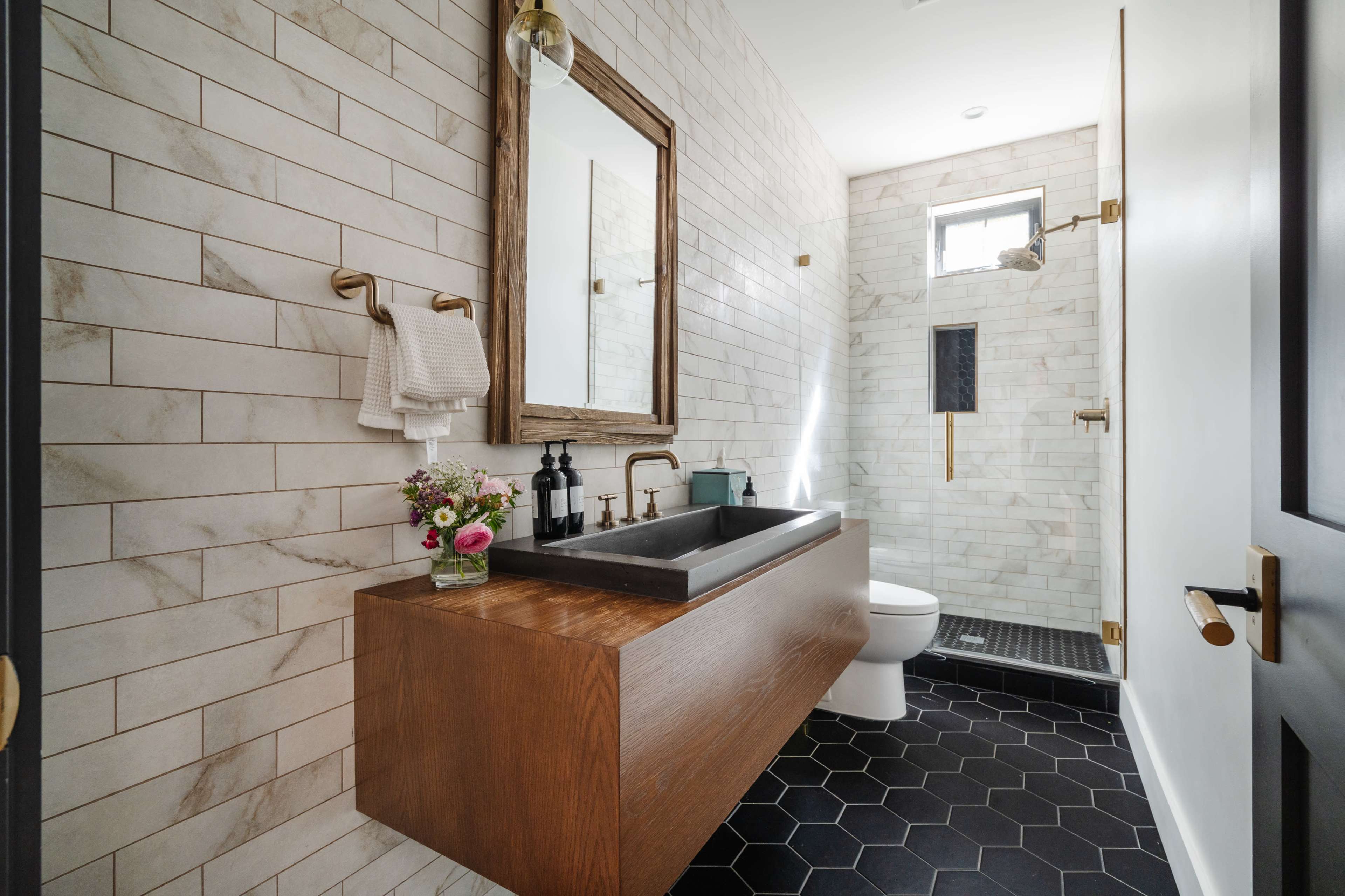 A modern bathroom features a wall-mounted sink with a wooden cabinet, a large mirror, and a hexagonal tile floor.
