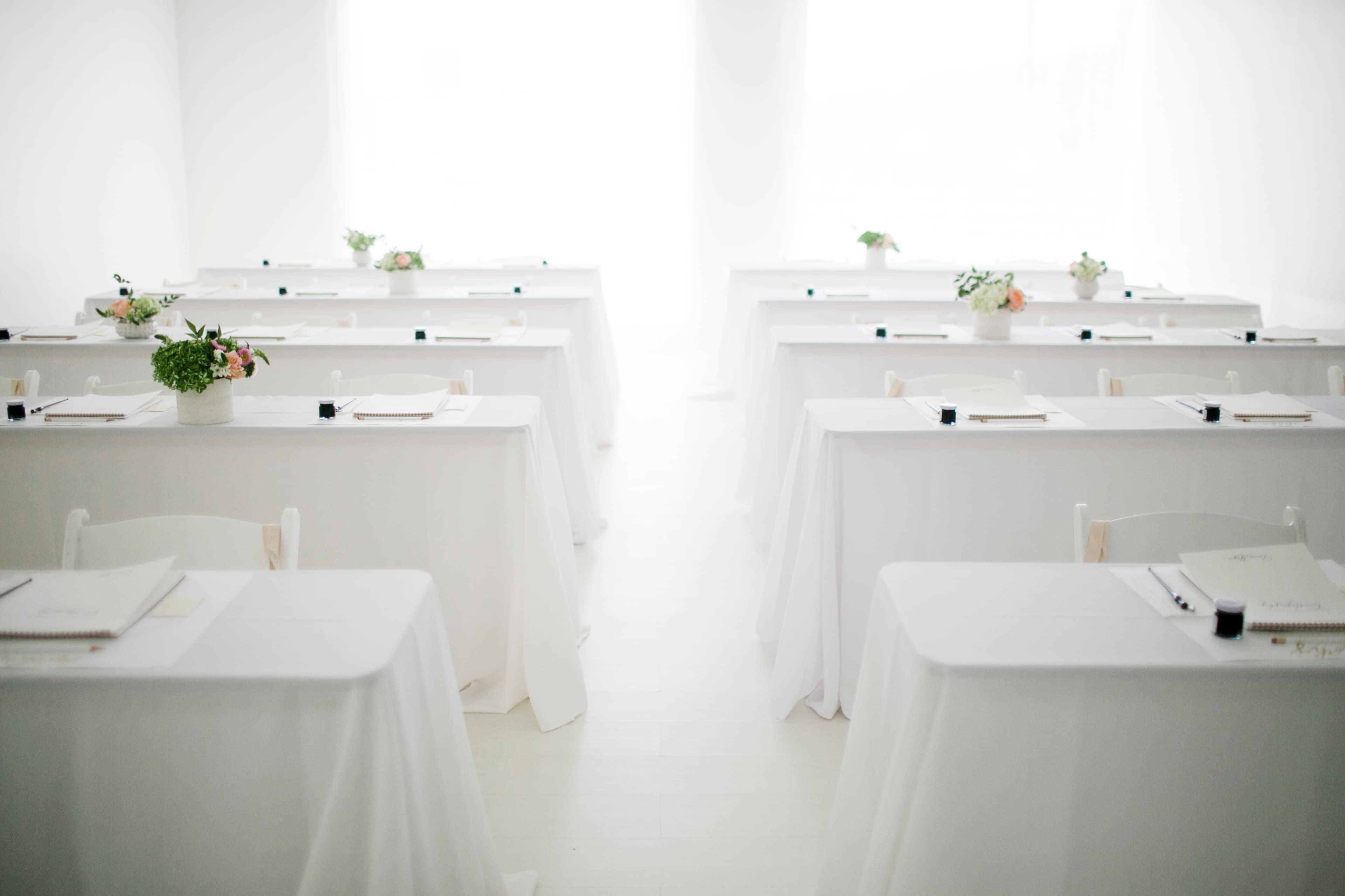 A bright, minimalistic room set up for a seminar or workshop, featuring neatly arranged tables with white tablecloths and floral centerpieces.