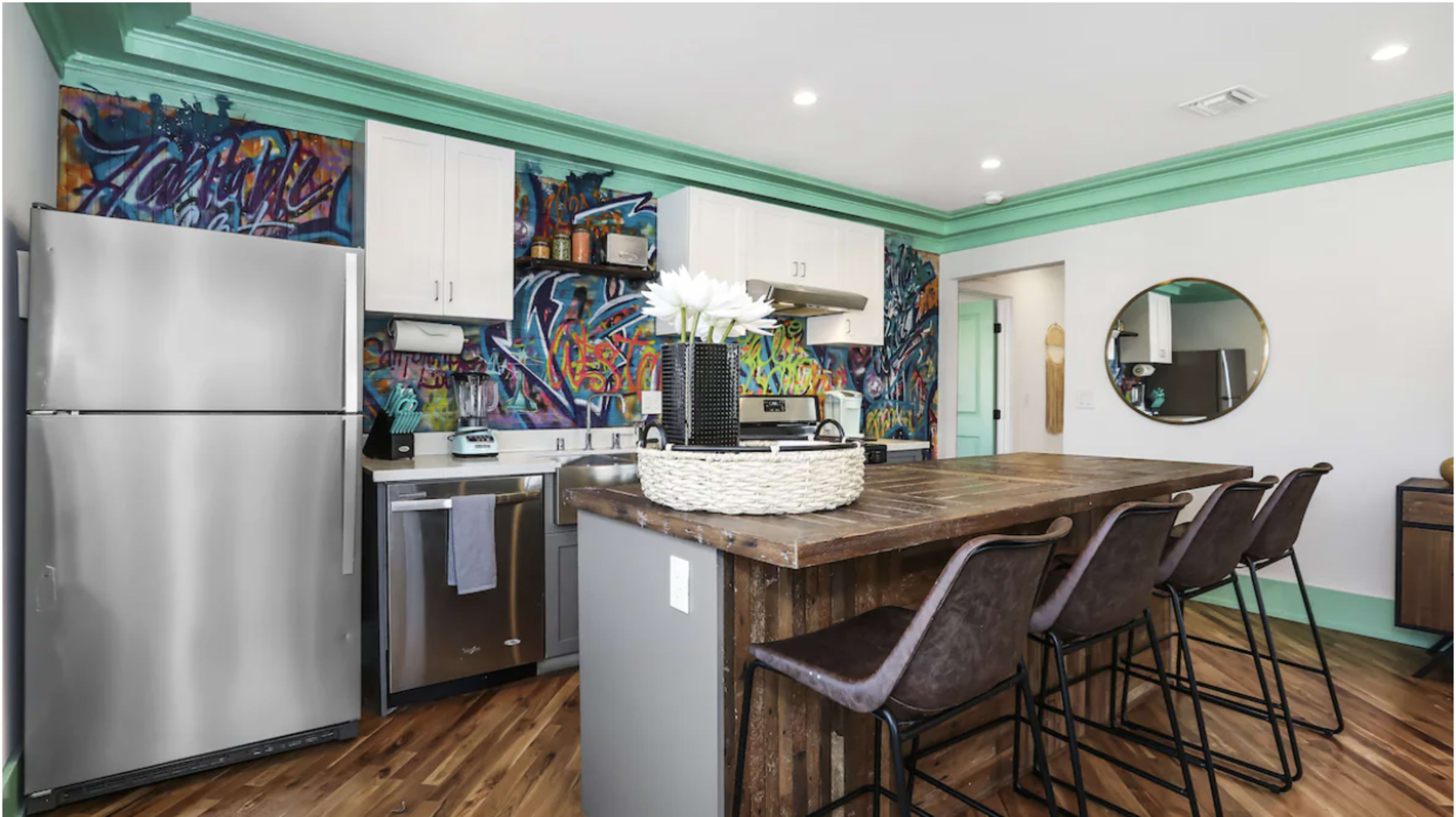 The image shows a modern kitchen with a vibrant, graffiti-style wall mural, stainless steel appliances, and a wooden island with bar stools.