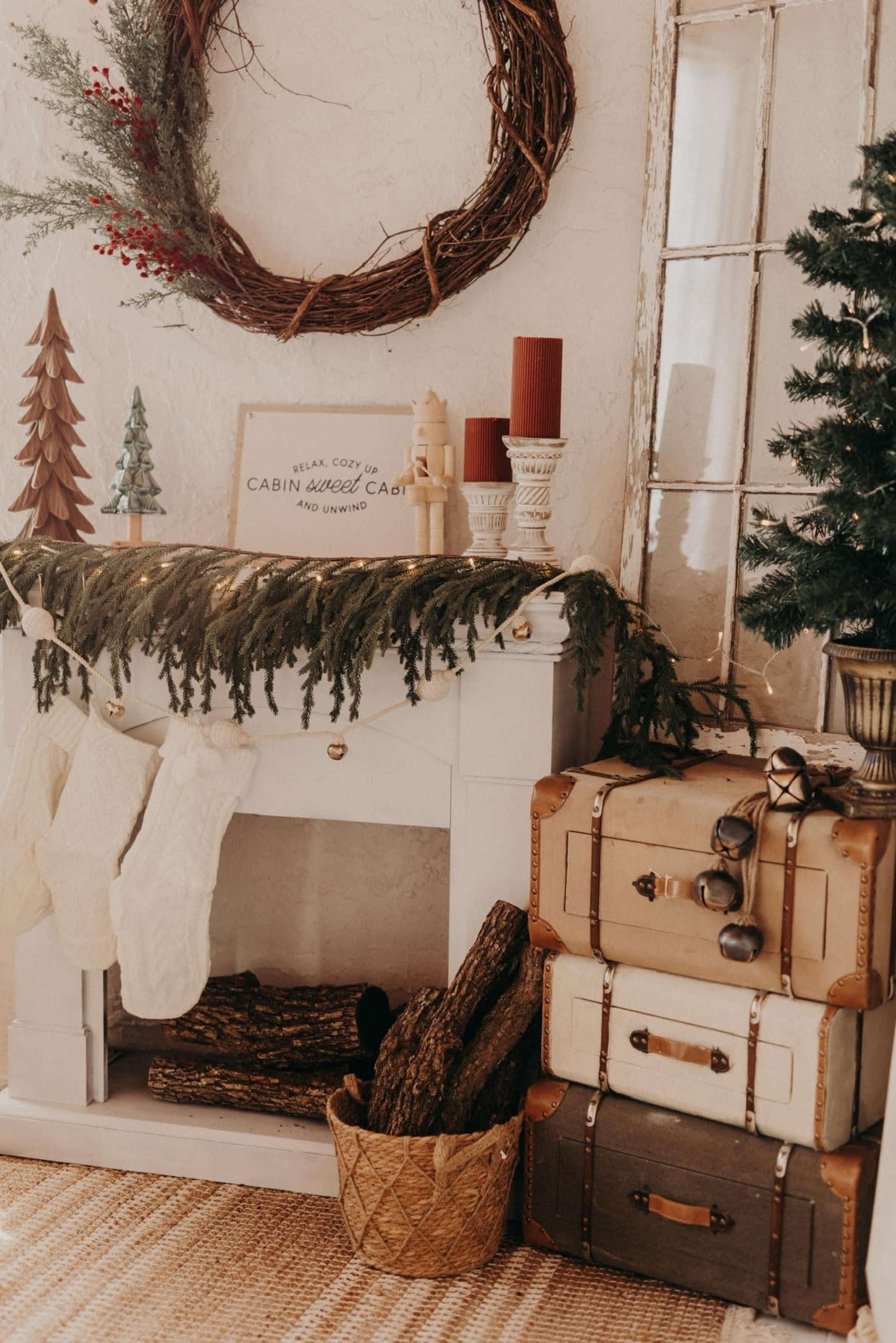 A decorated mantel features a wreath, candles, stockings, and a stack of vintage suitcases alongside a small Christmas tree.