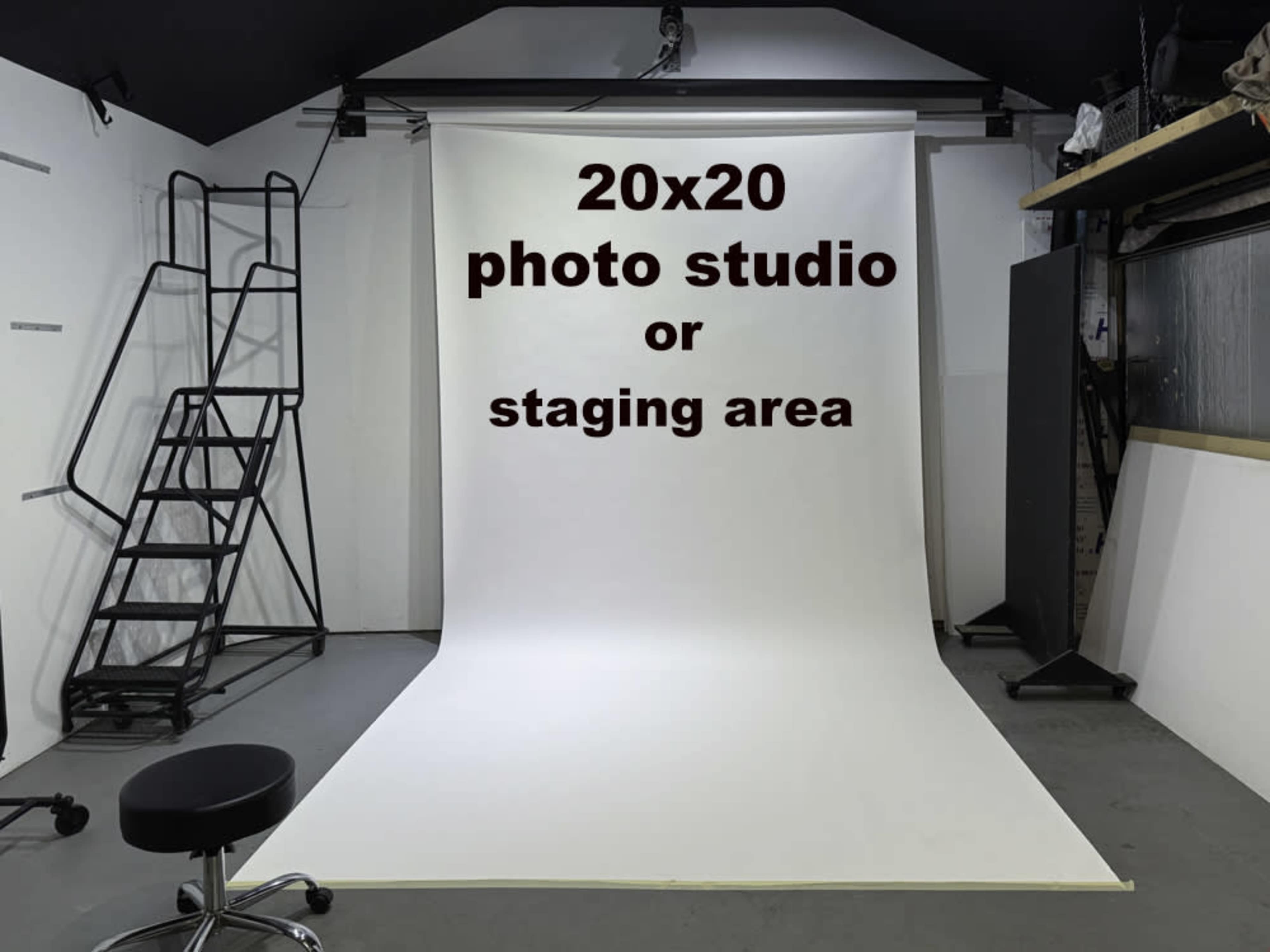 The image shows a simple, empty photo studio with a large white backdrop, a stool, and a ladder in the corner.