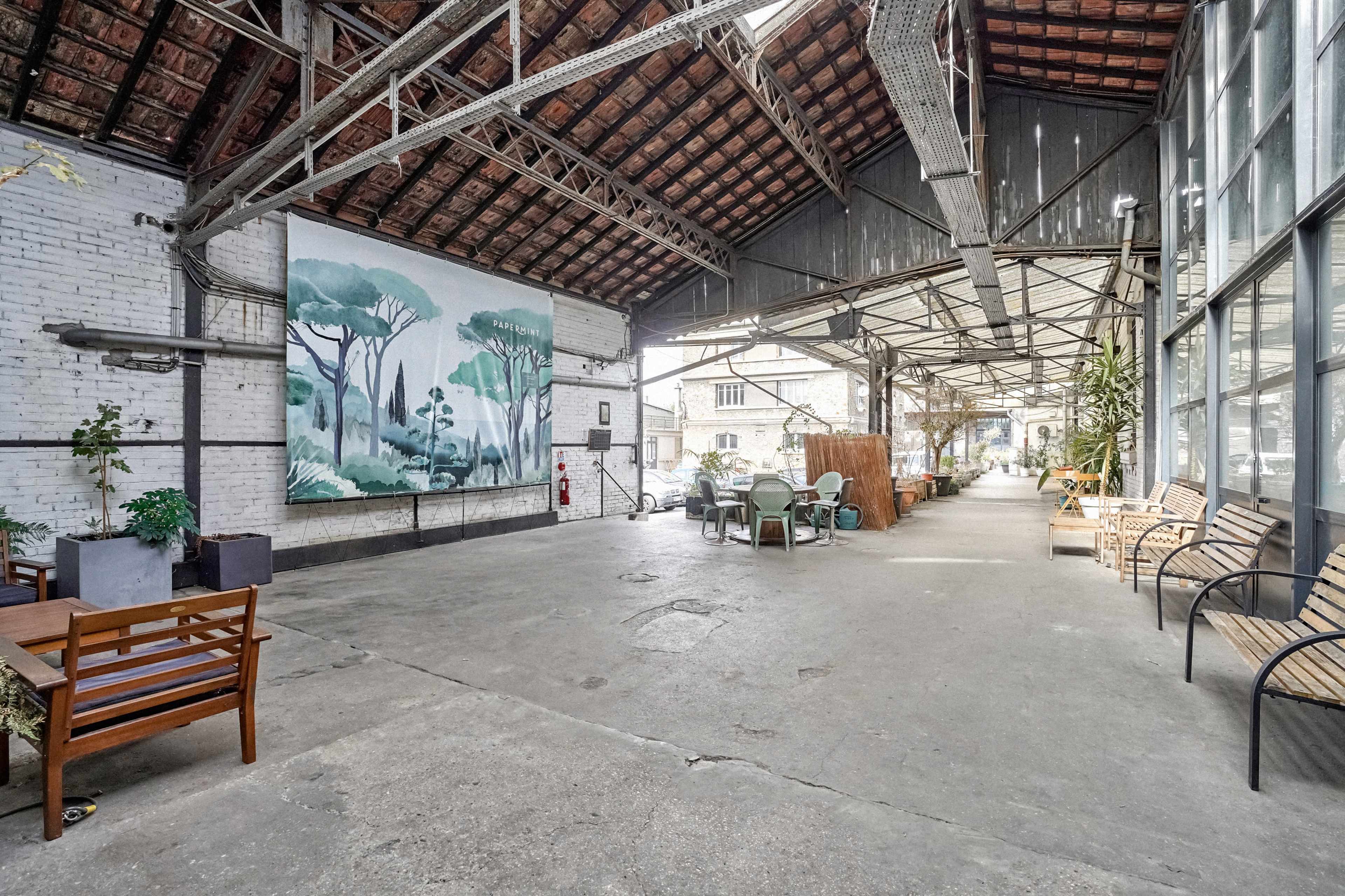 The image depicts a spacious, industrial-style corridor with a mural of trees on one wall and several seating areas along the sides.