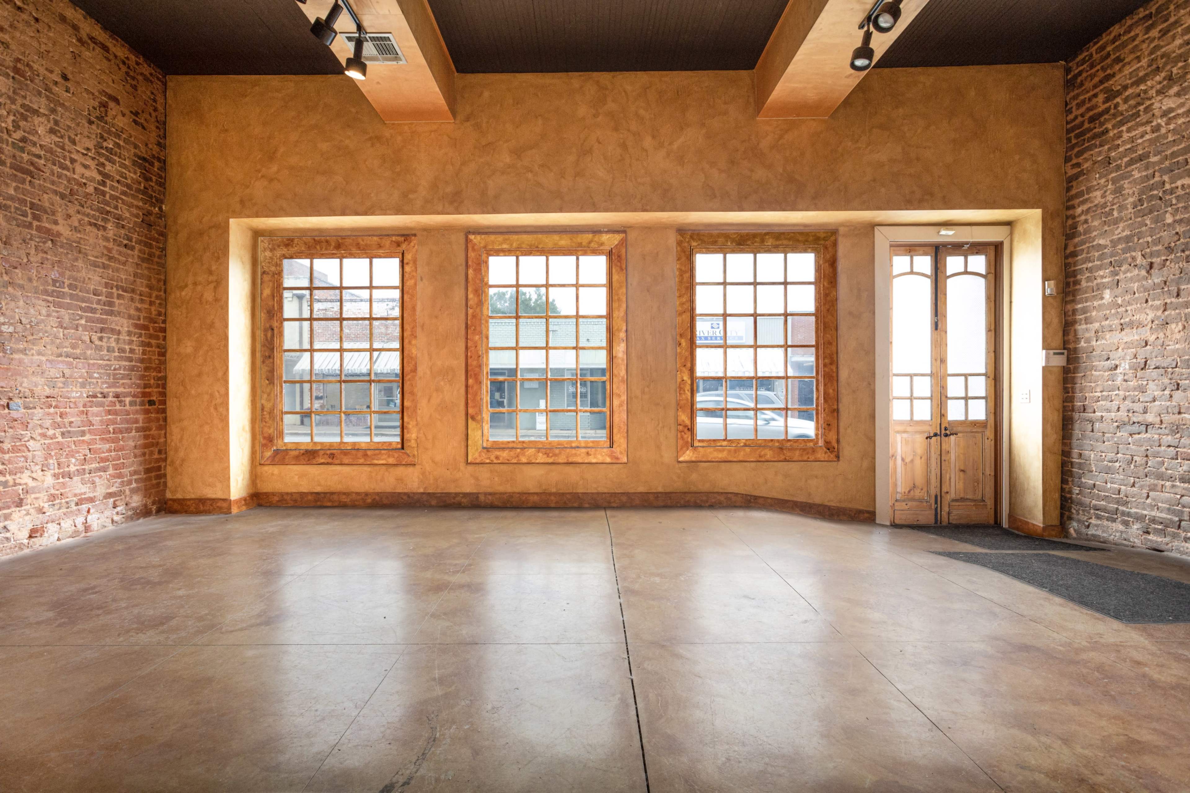The image shows an empty room with exposed brick walls, large windows, and a wooden door leading outside.