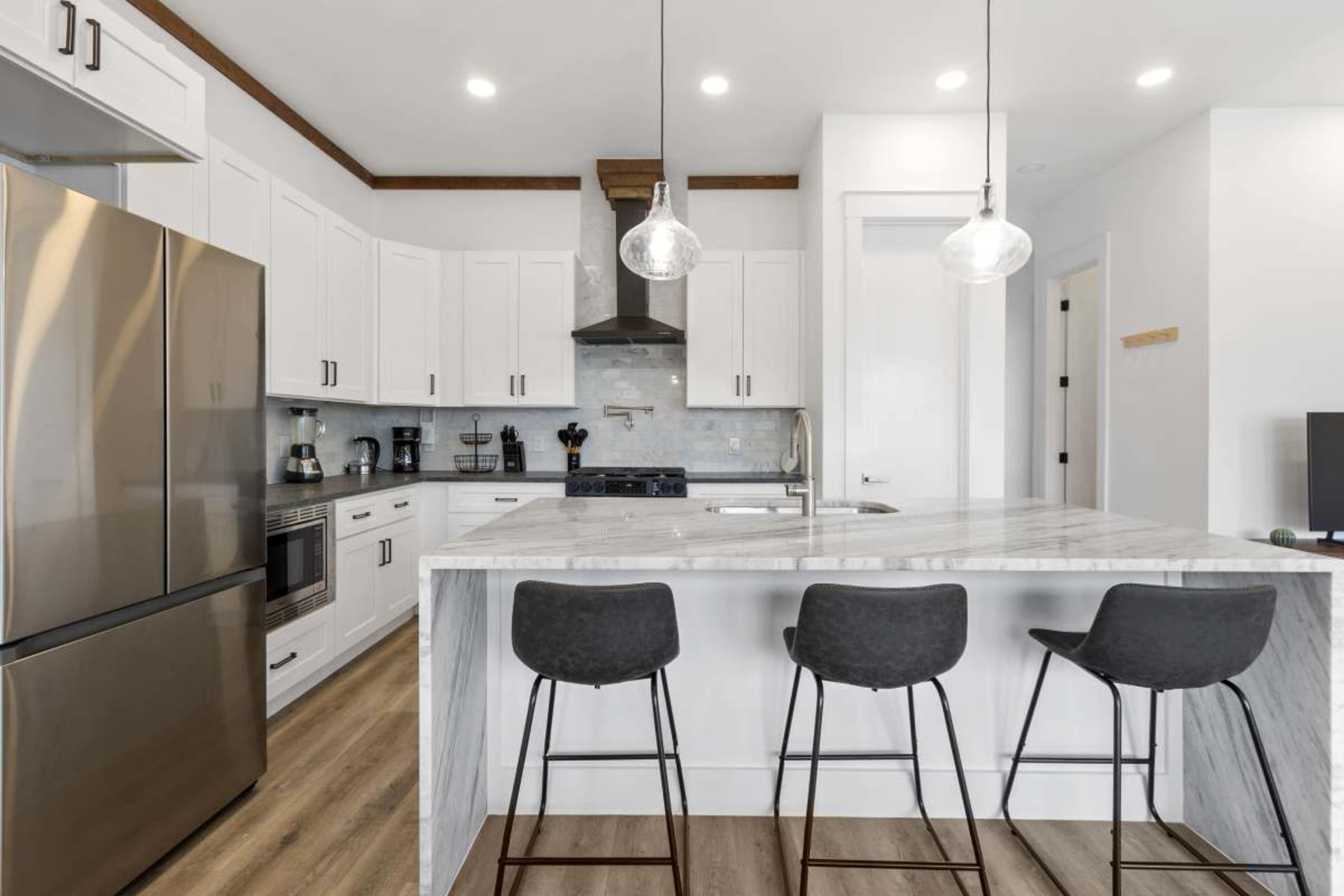 A modern kitchen features stainless steel appliances, white cabinets, a marble island with three black bar stools, and pendant lighting.