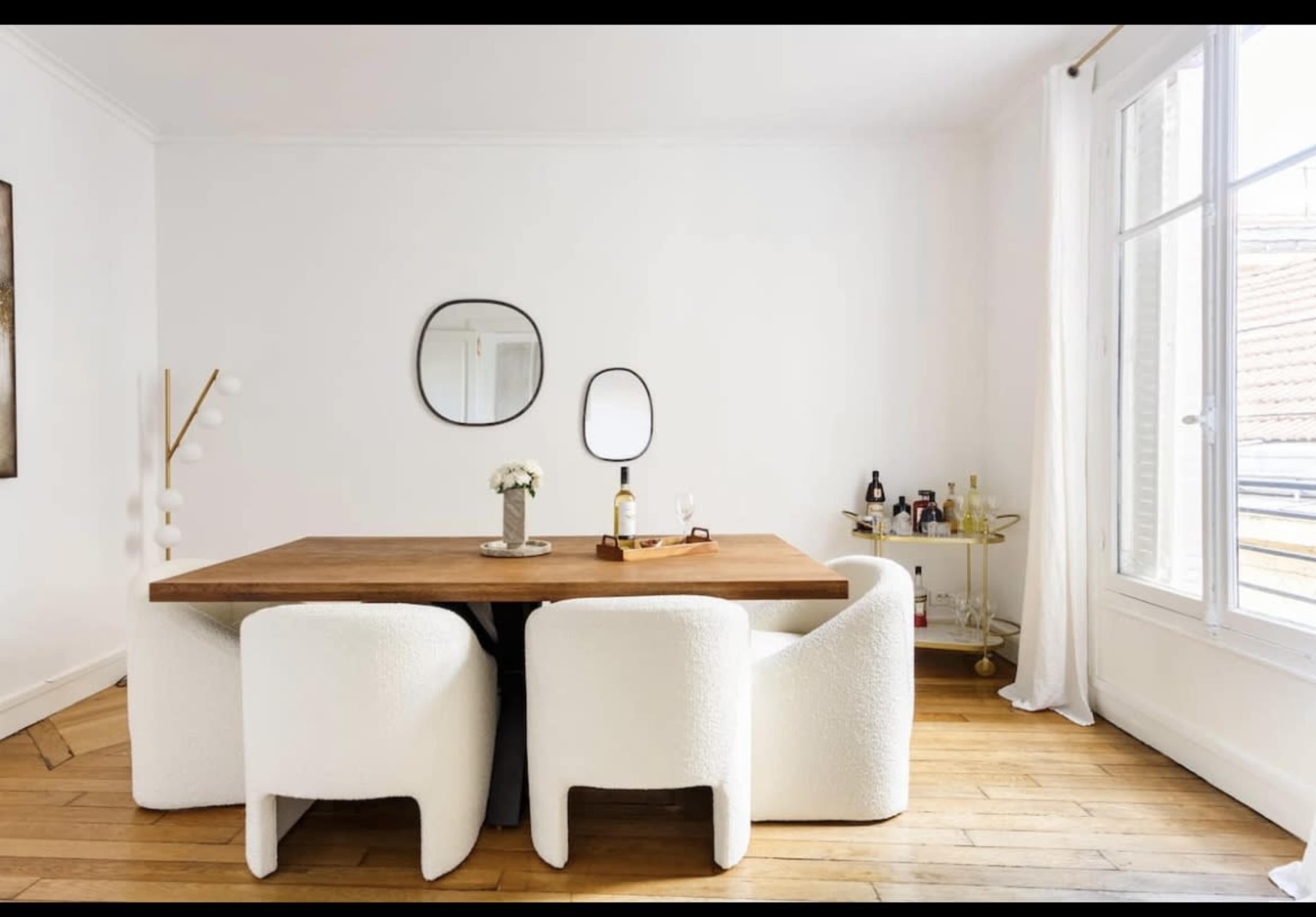 A wooden dining table is surrounded by four white, cushioned chairs in a bright room with large windows and minimalist decor.