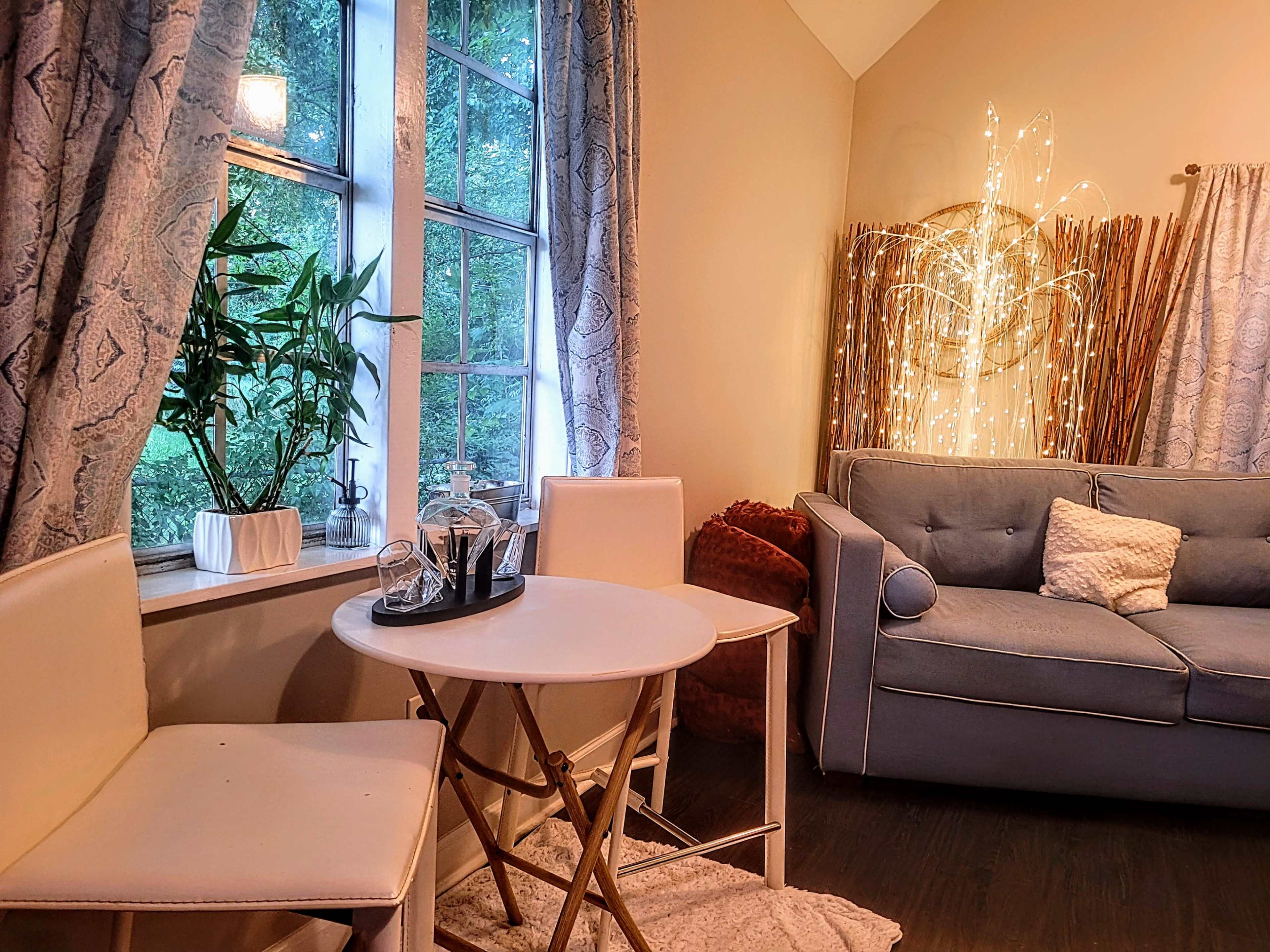 A small dining area features a round table with two chairs beside a window, a cozy couch, and decorative lighting in the background.