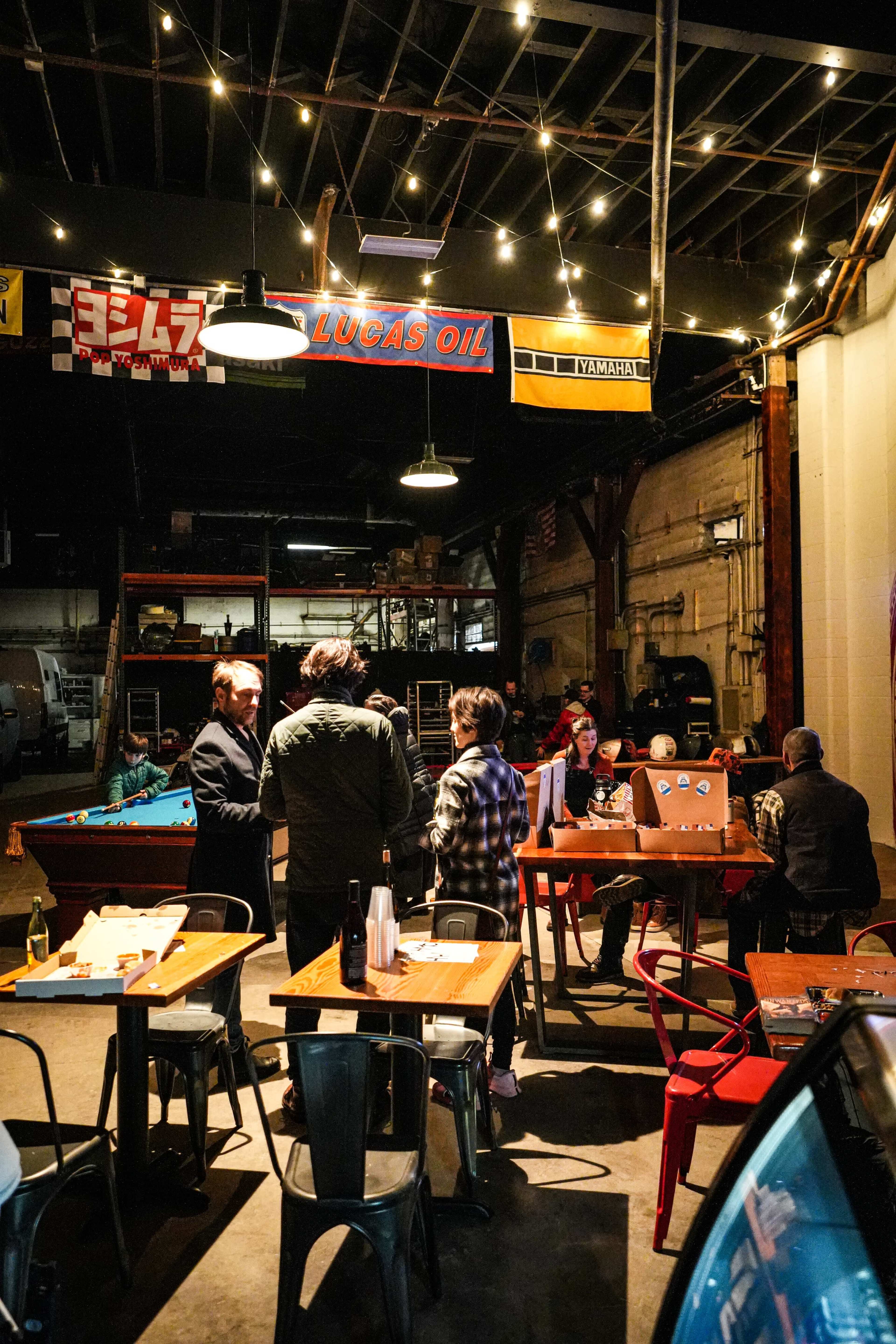 A group of people stands conversing in a dimly lit industrial space with pool tables and string lights overhead.