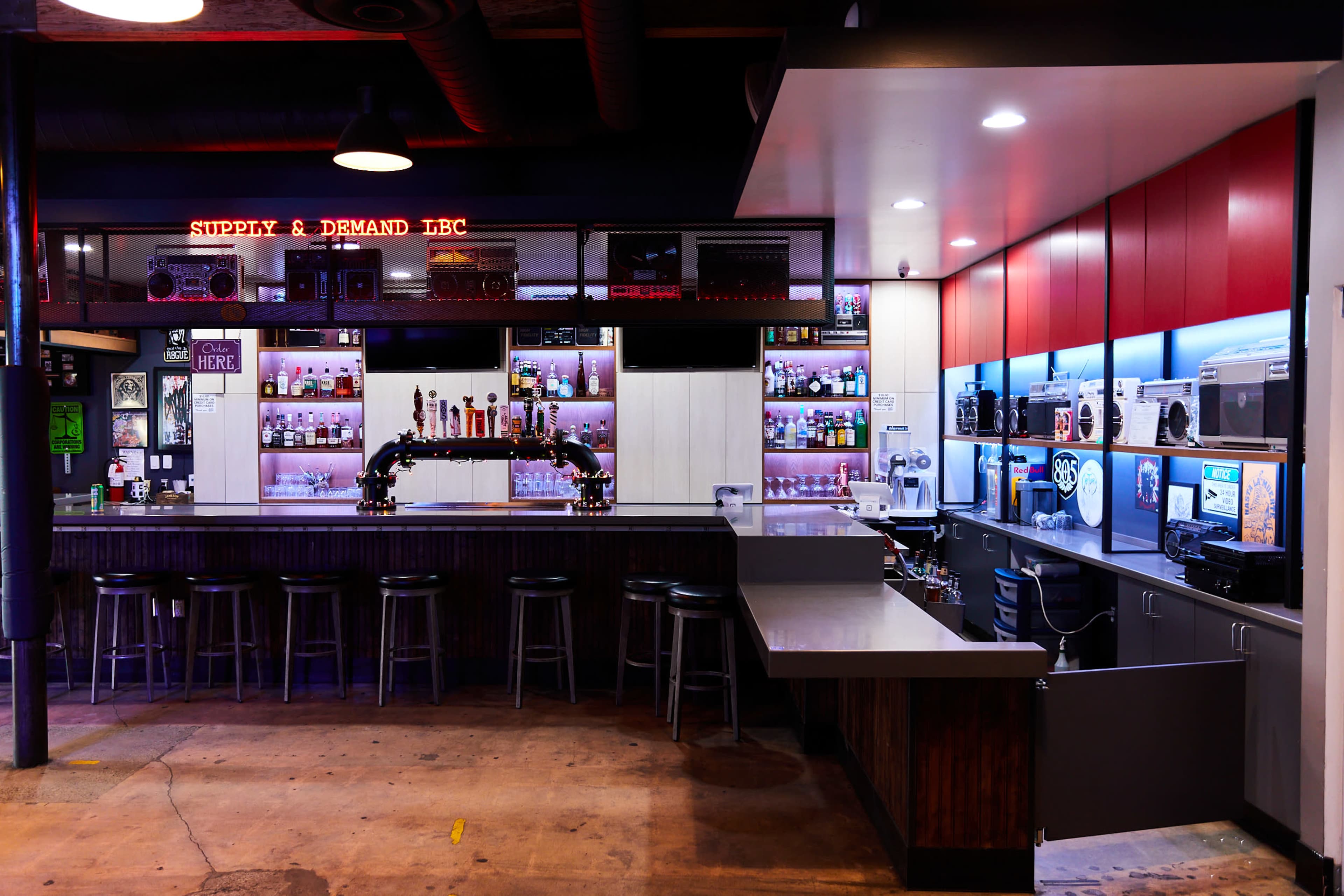 A modern bar with a Long counter, shelves of various bottles, and seating areas, all illuminated by ambient lighting.