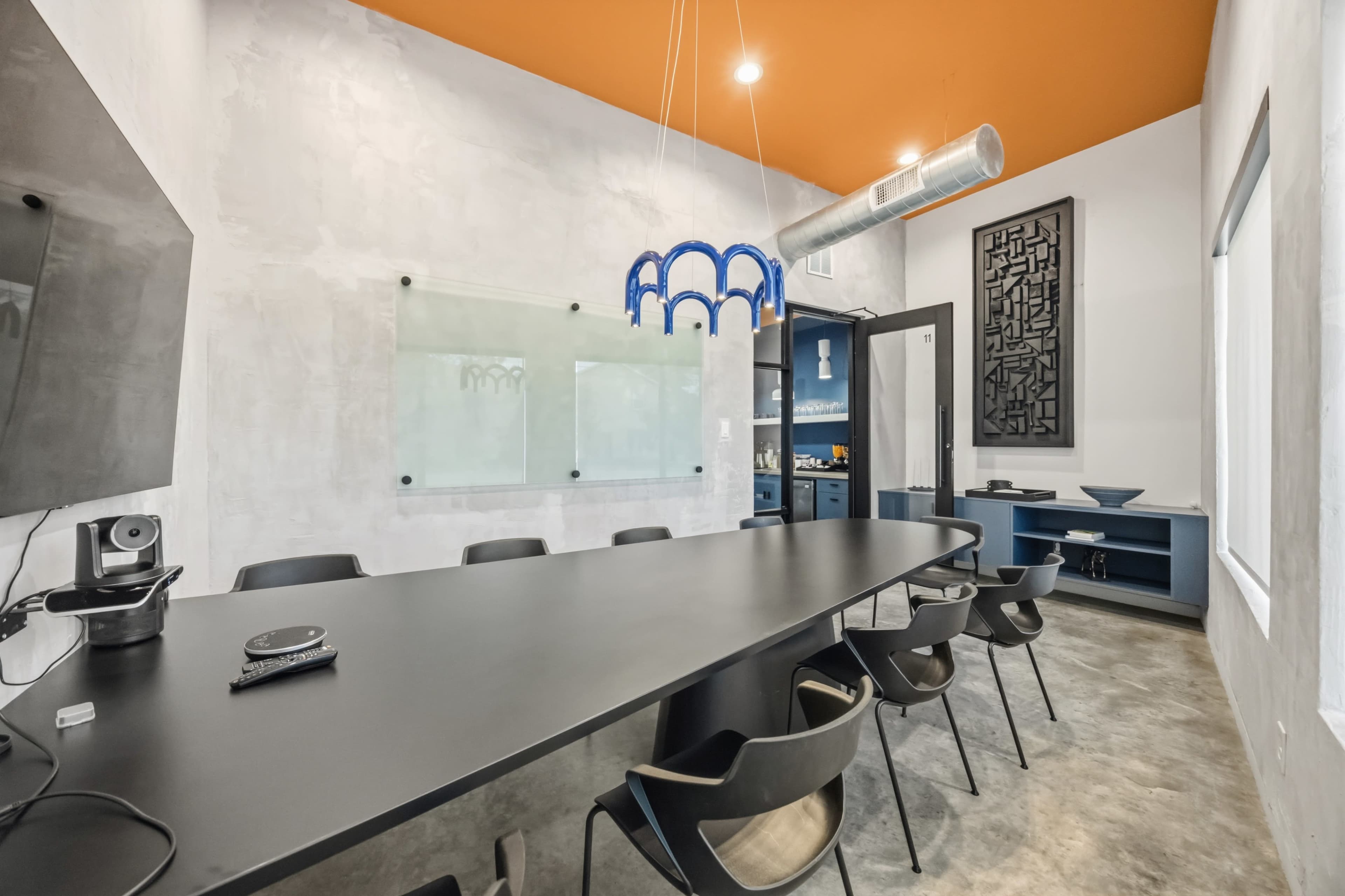 A modern conference room features a long black table surrounded by black chairs, with a concrete wall and an orange ceiling.