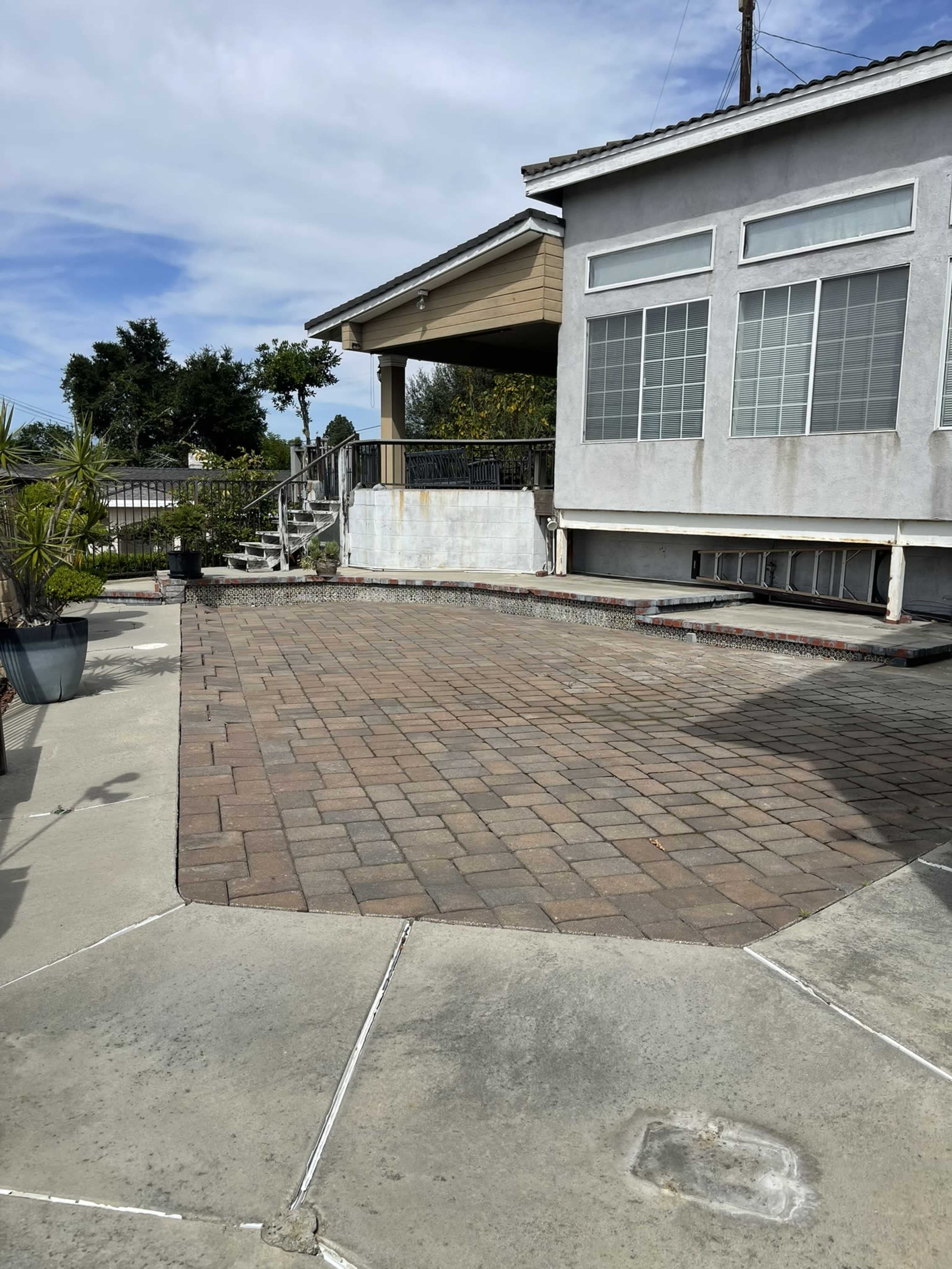 The image shows a patio area made of paver stones adjacent to a house with large windows and a staircase leading up to a covered porch.