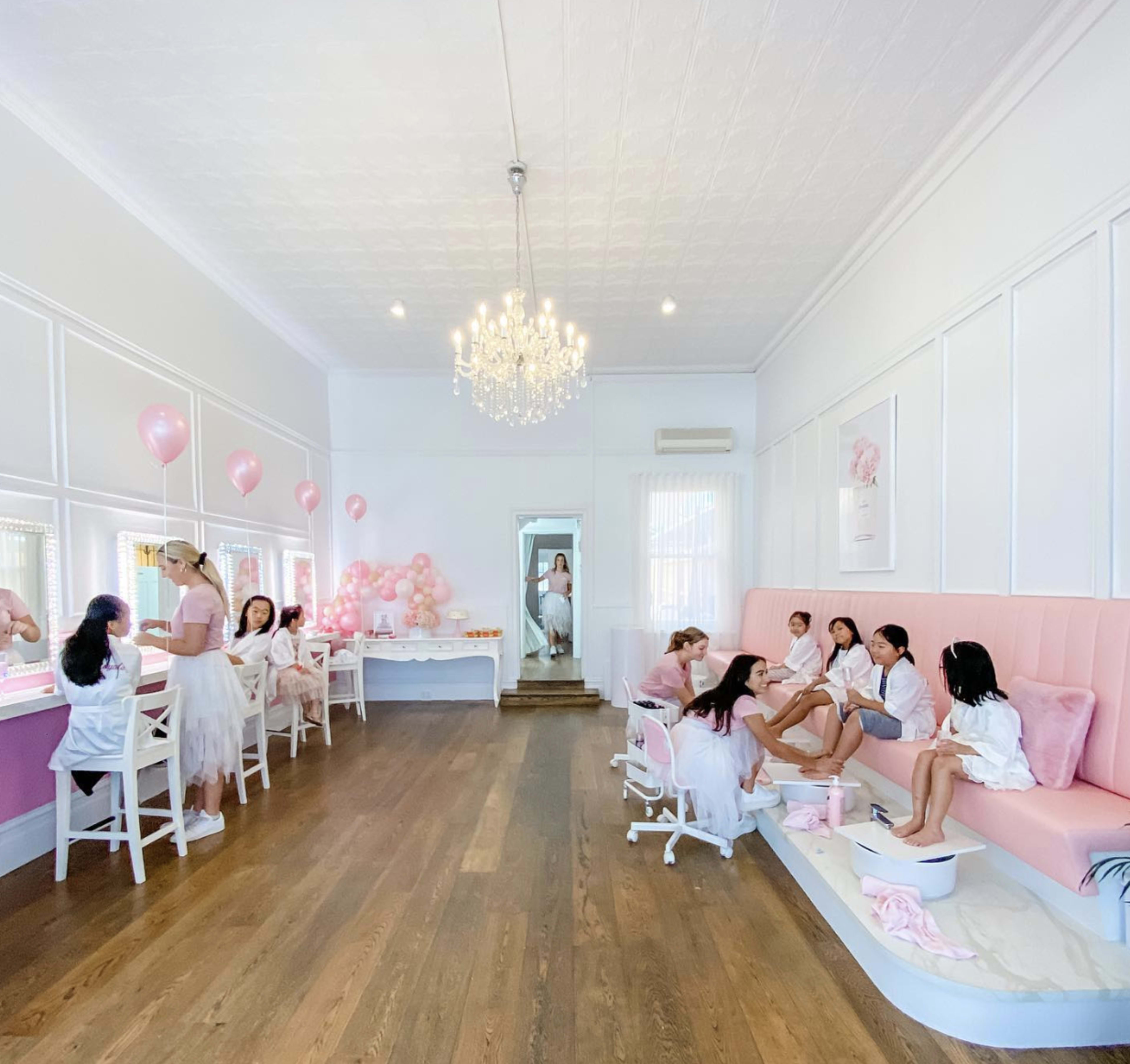 A bright salon features several women and girls receiving pedicures while others enjoy treatments in a pastel pink-themed space.