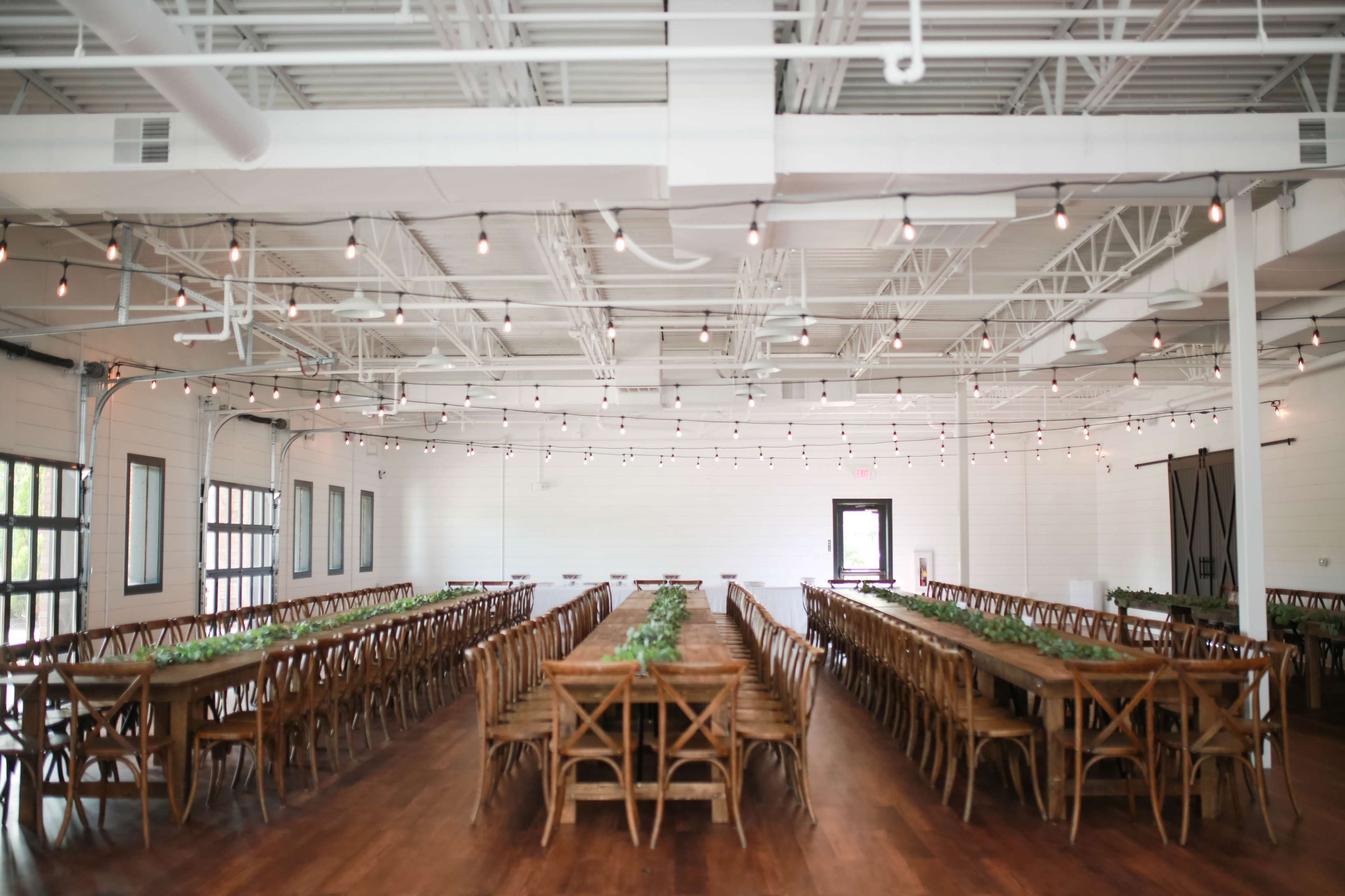 A spacious, well-lit event hall features long wooden tables arranged in parallel rows, with string lights hanging overhead.