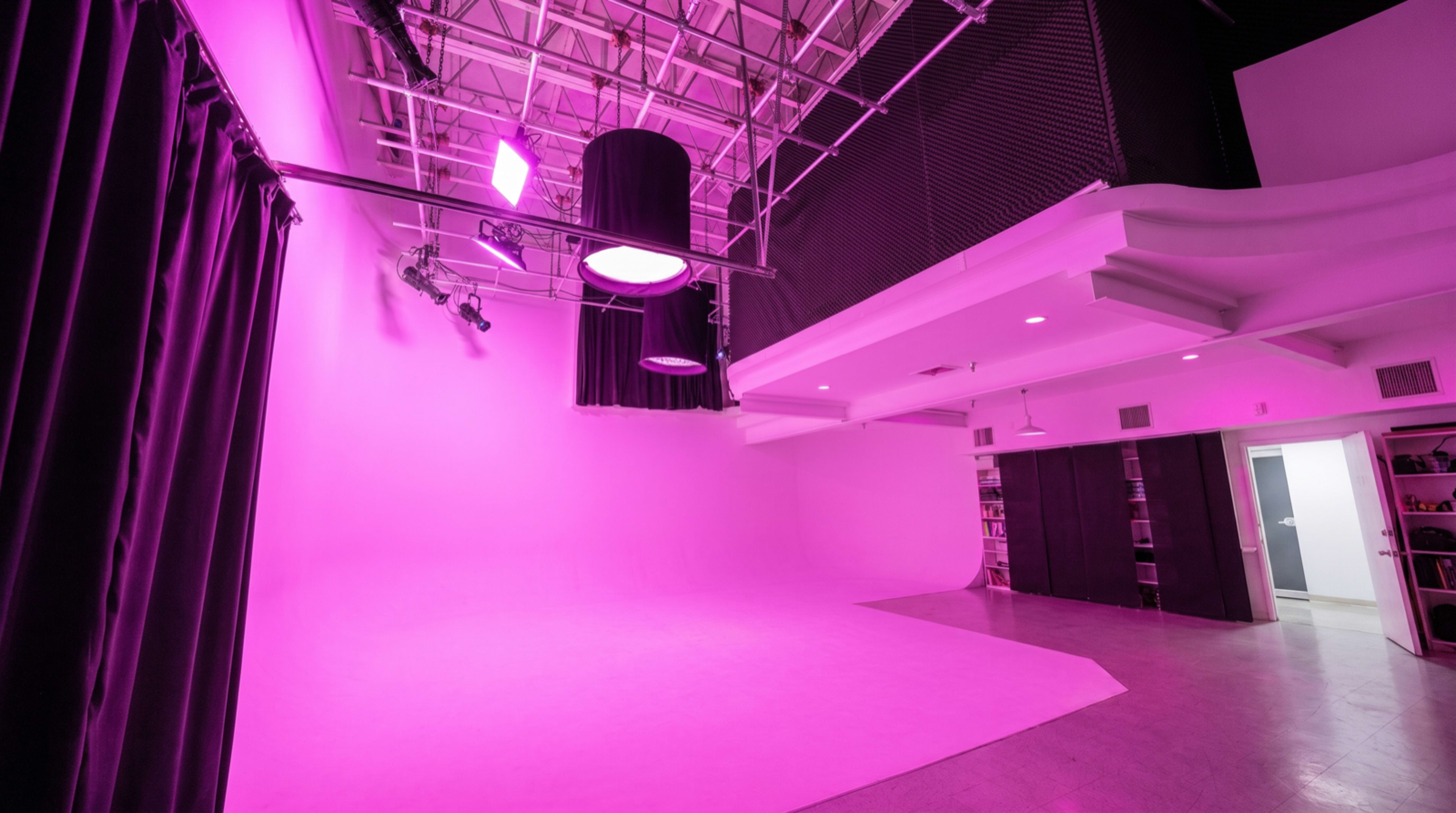 The image shows a brightly lit photography studio with pink lighting, a large backdrop area, and black curtains along one side.