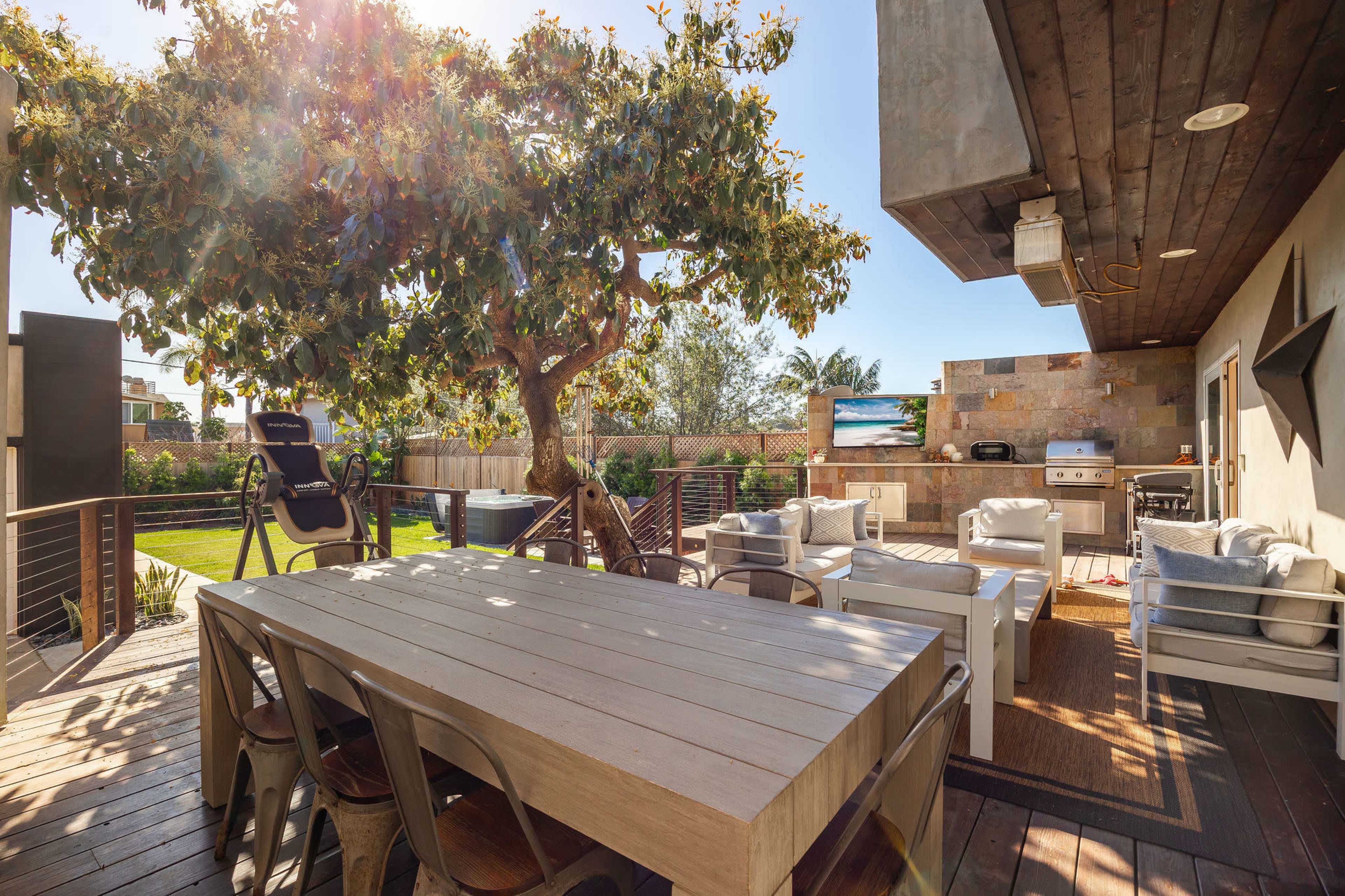 A spacious outdoor patio features a large wooden dining table, comfortable seating, a tree, and a grill under a clear blue sky.