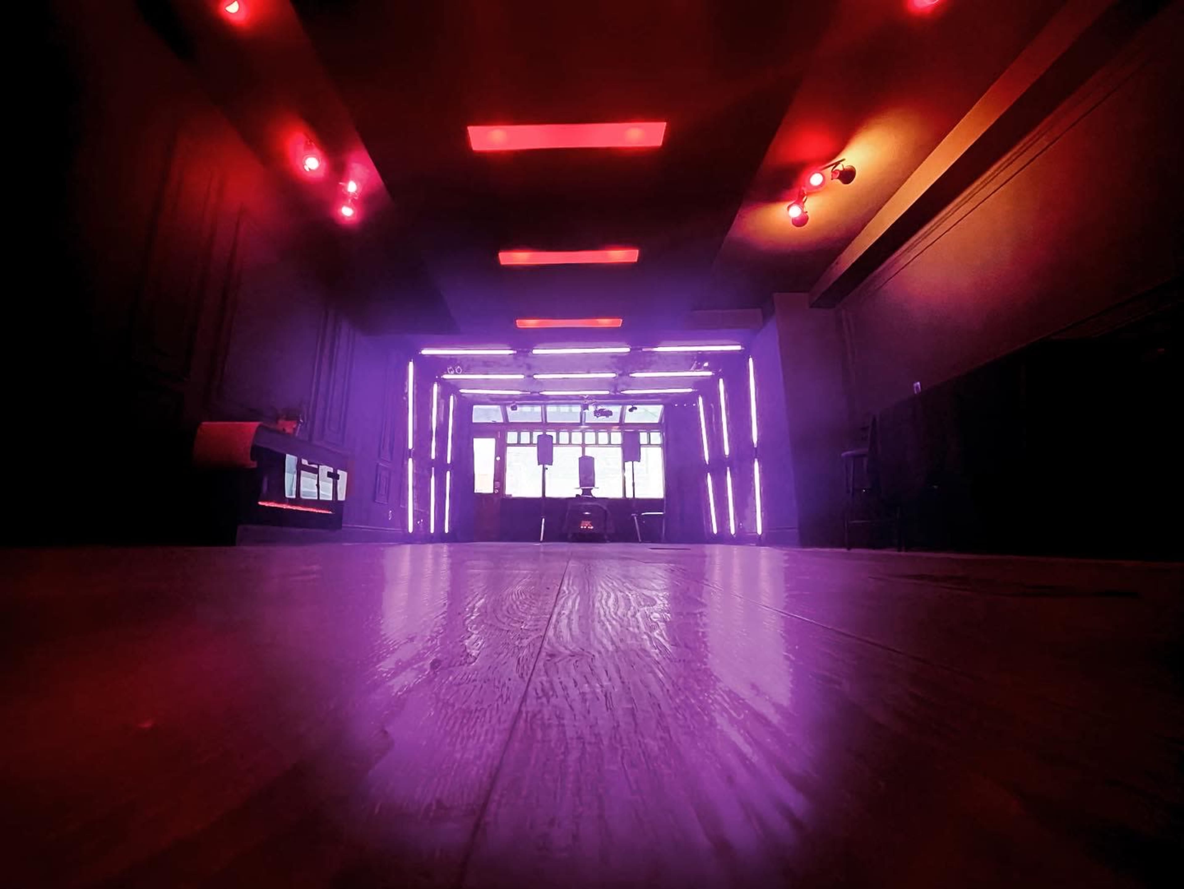 A dimly lit room with purple and red lighting, featuring a wooden floor and large windows in the background.