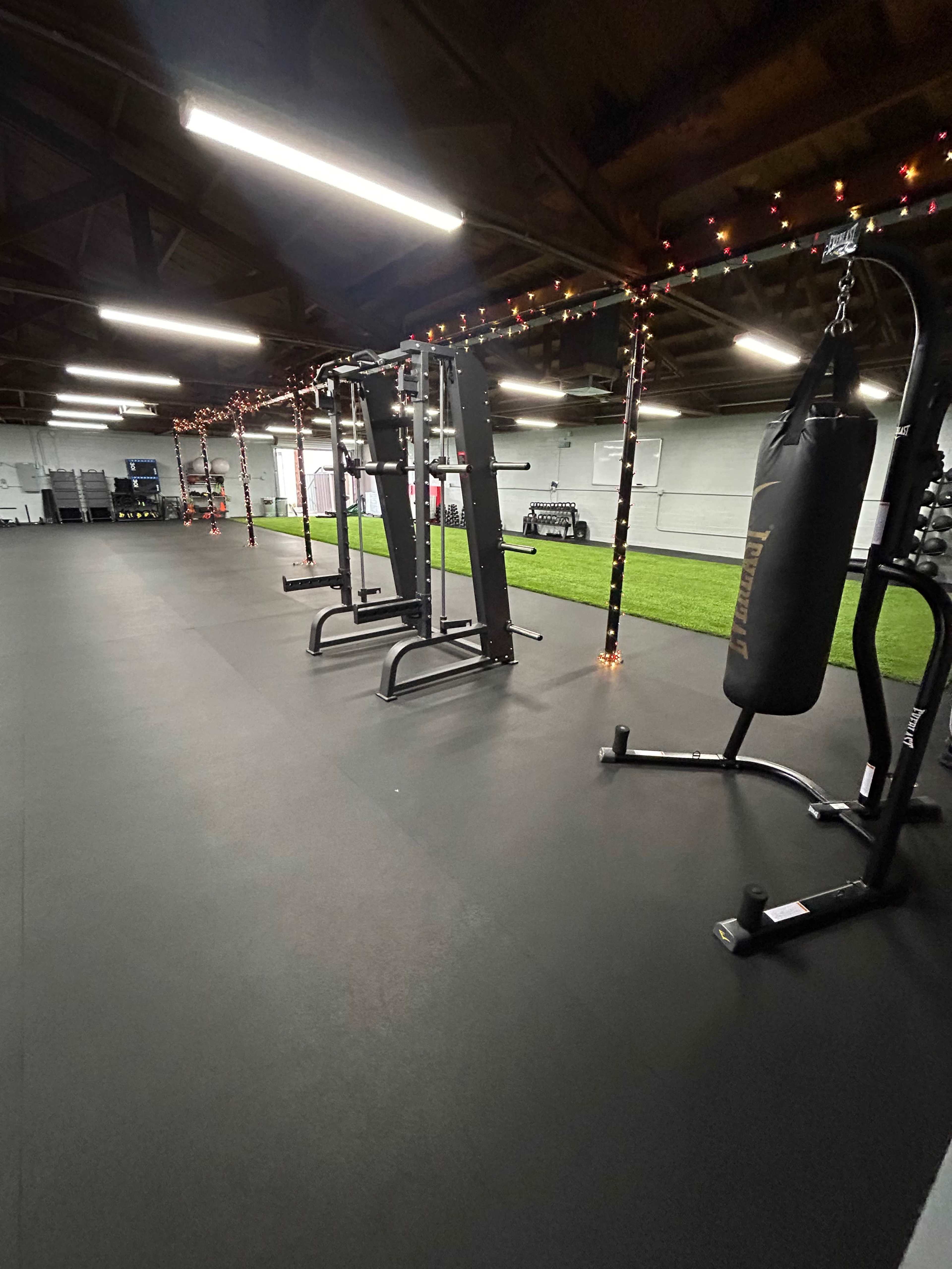 A fitness room equipped with exercise machines, a punching bag, and a turf area, illuminated by overhead lights and decorated with string lights.