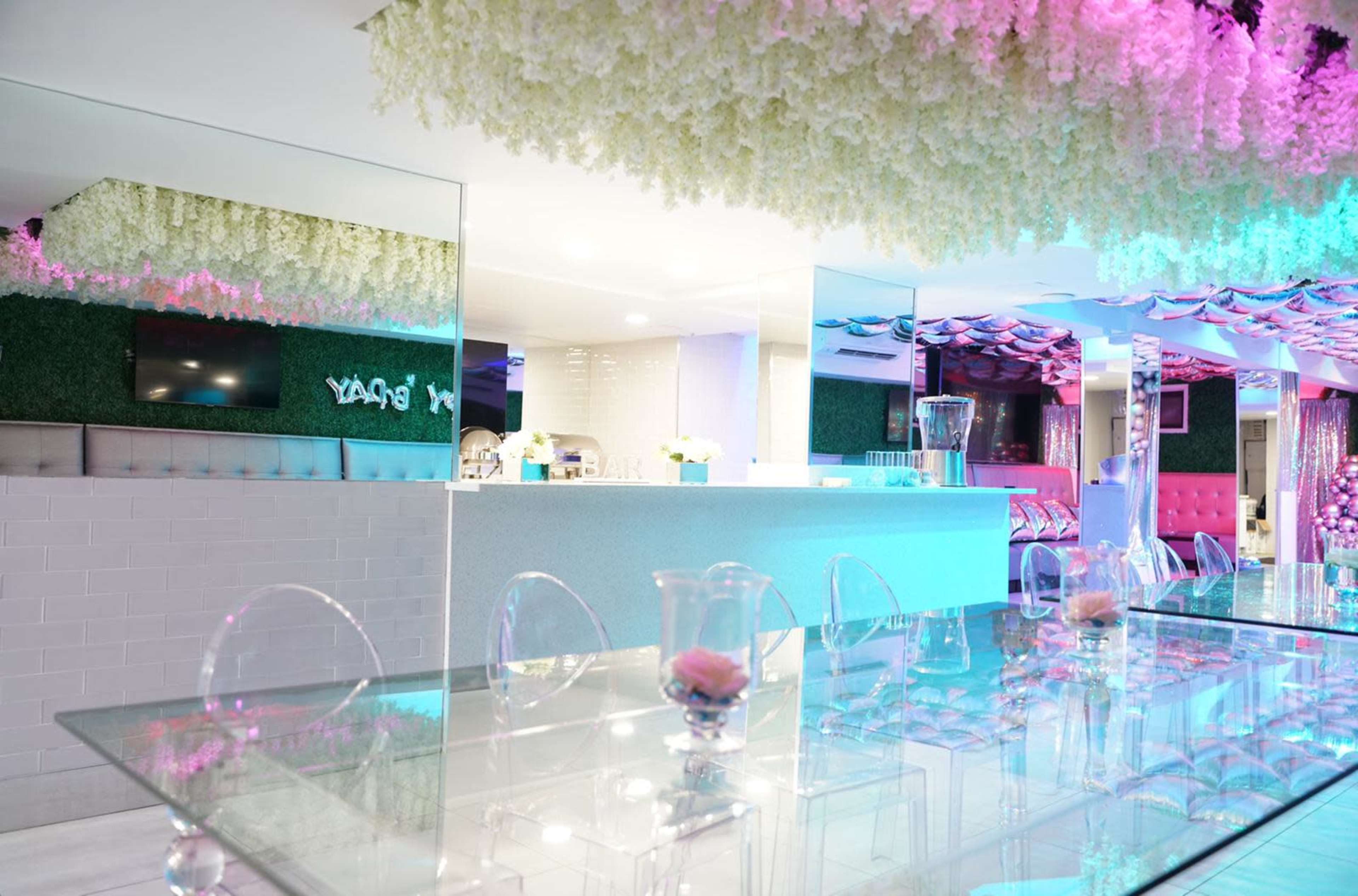 A modern, brightly lit interior of a restaurant or event space with floral decorations hanging from the ceiling and a glass dining table at the center.