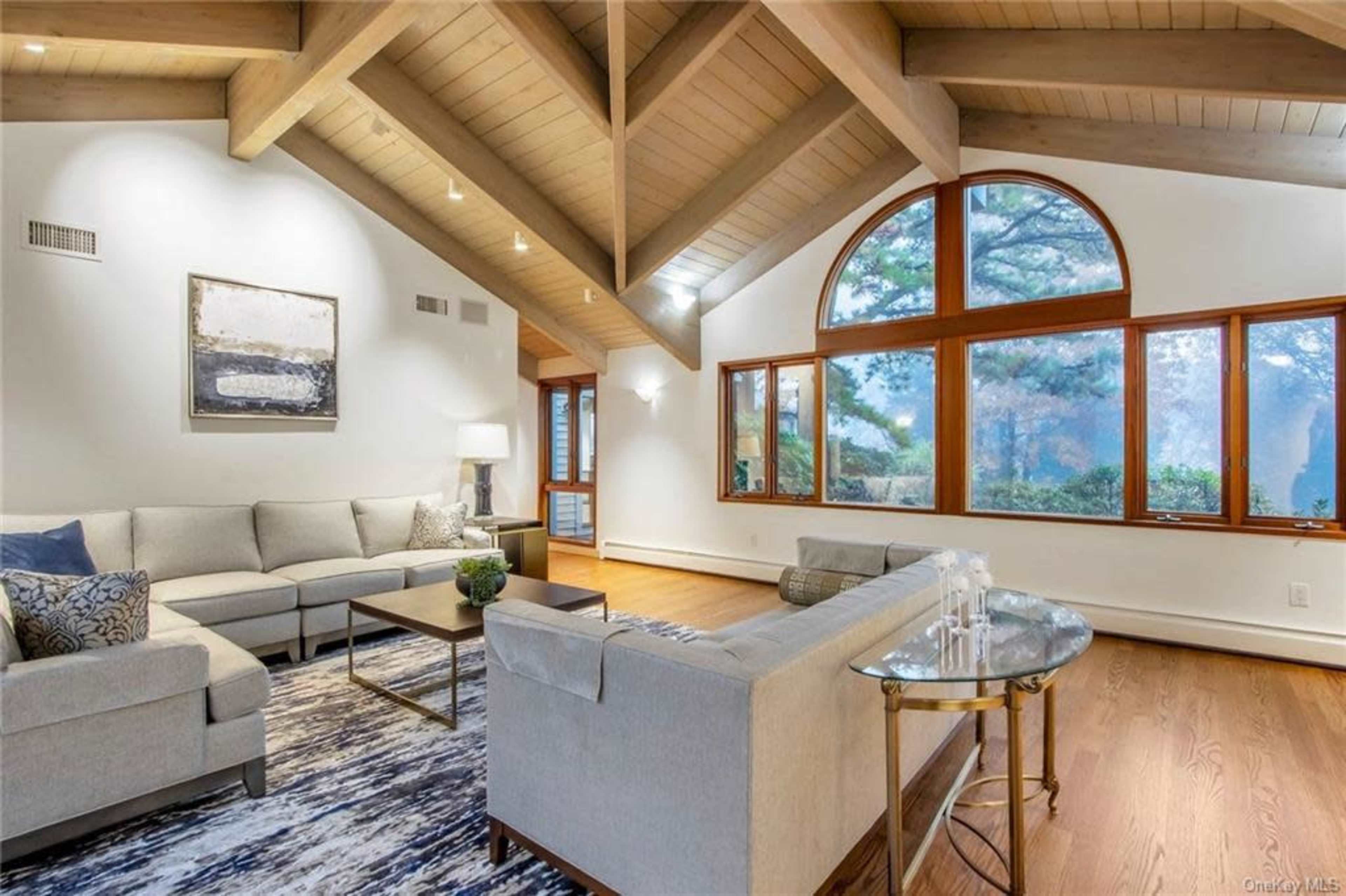 The image shows a modern living room featuring a light gray sectional sofa, large windows with wooden frames, and a vaulted ceiling with exposed beams.