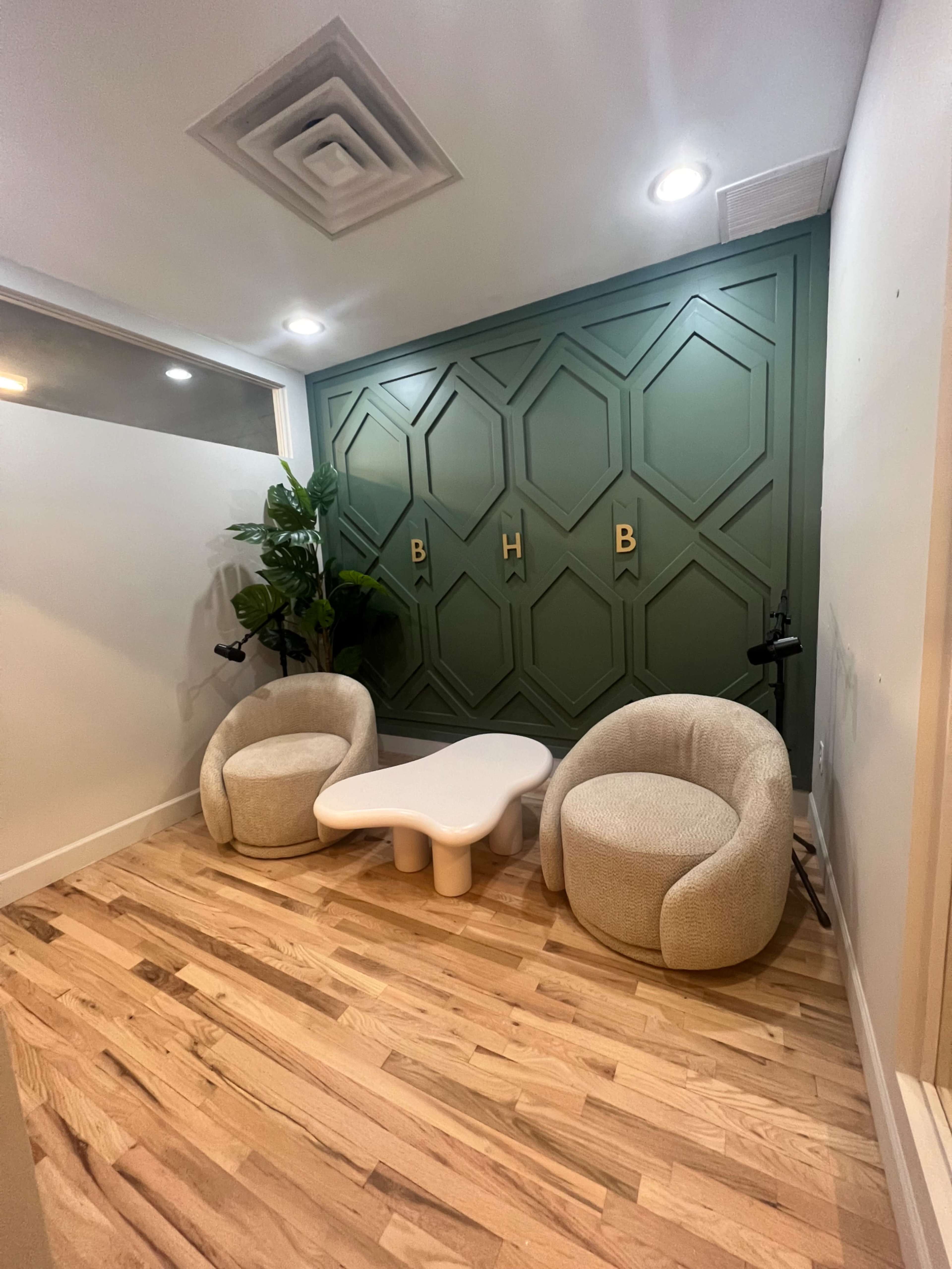 A small lounge area featuring two rounded chairs, a white coffee table, and a decorative green accent wall with wooden flooring.