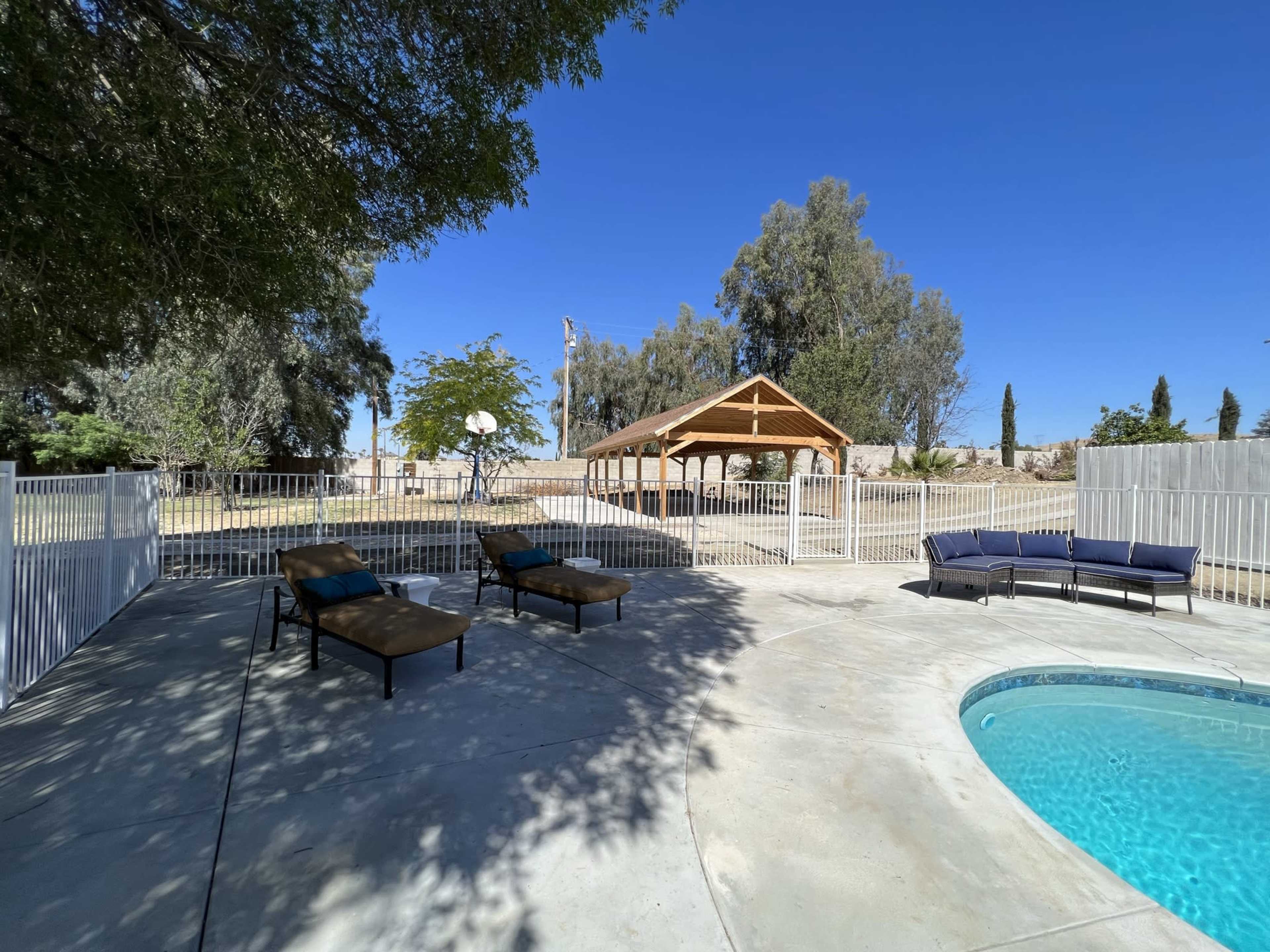 A backyard with a swimming pool, lounge chairs, a covered pavilion, and a basketball hoop in the distance, surrounded by trees.