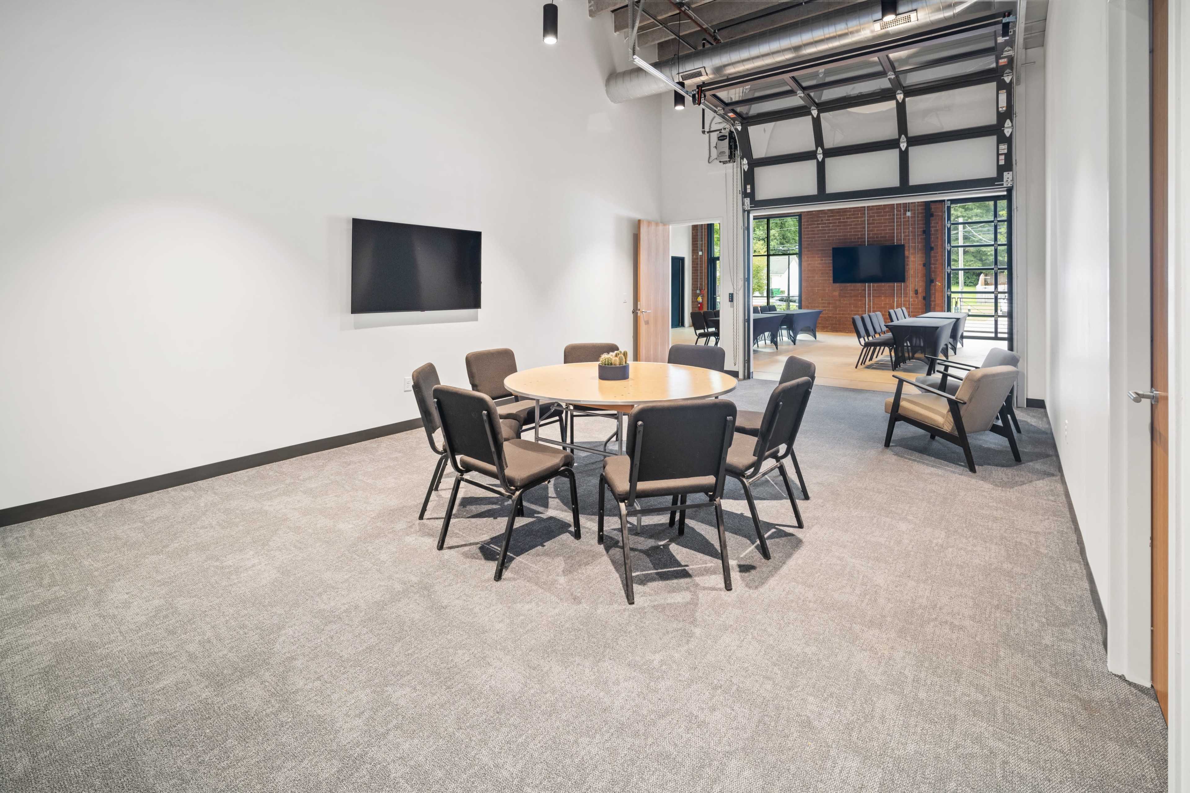 The image shows a modern conference room with a circular table surrounded by chairs, and a garage-style door leading to an outdoor area.