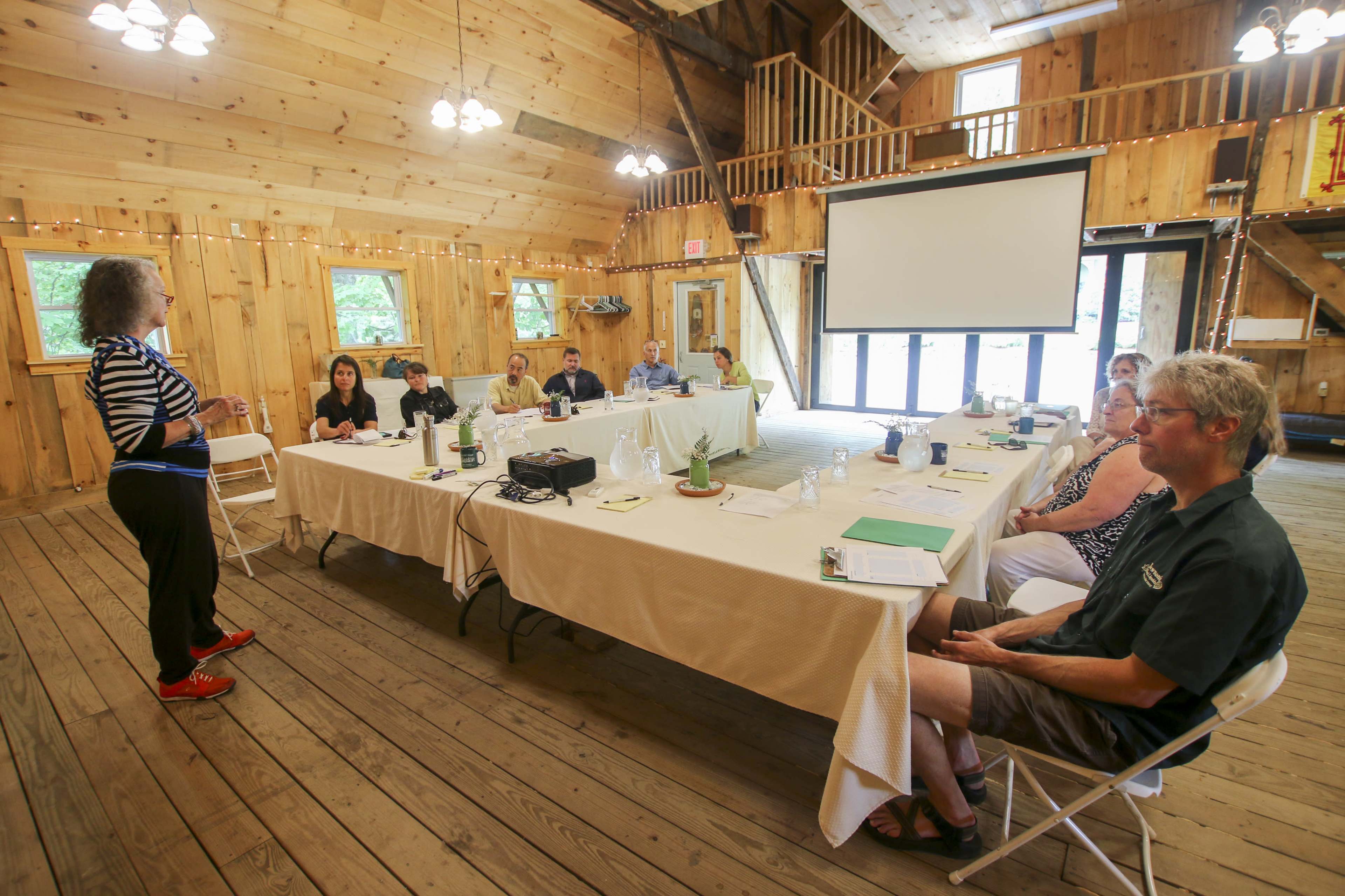 A group of people is seated at a long table in a wooden meeting room while a woman stands and speaks at the front.