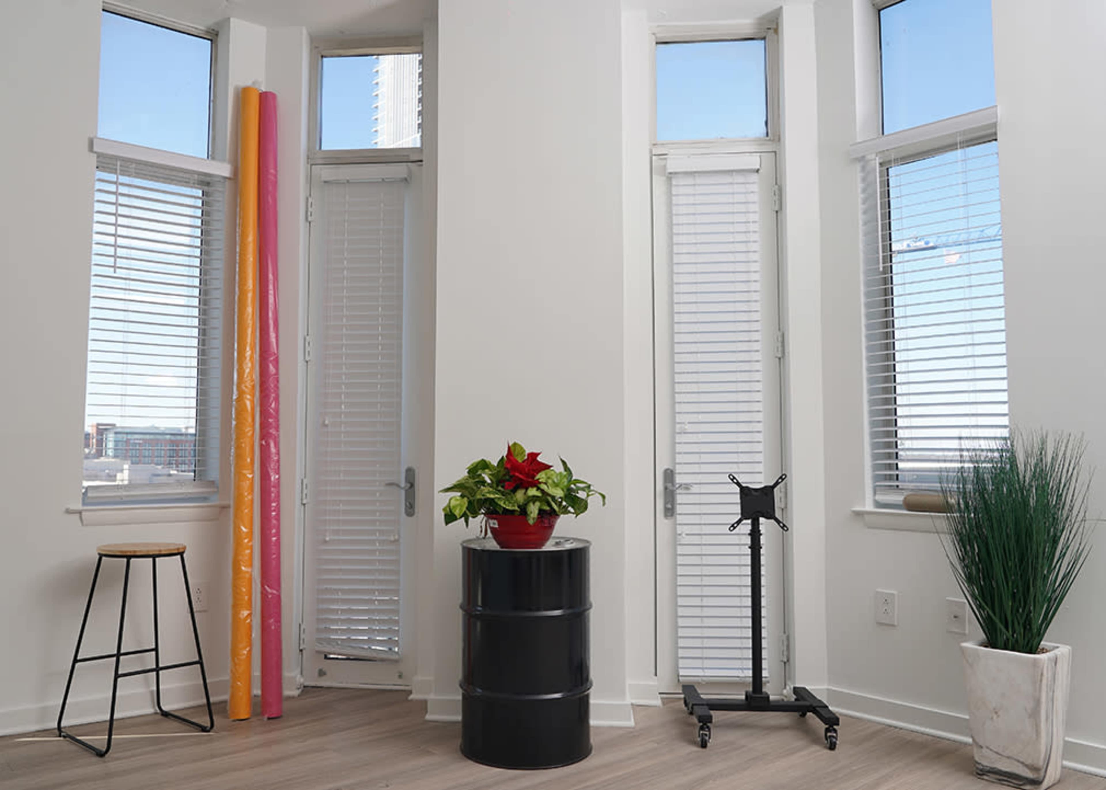 The image shows a bright room with large windows featuring blinds, a black barrel serving as a table, a stool, a potted plant, and rolls of colored paper leaning against the wall.