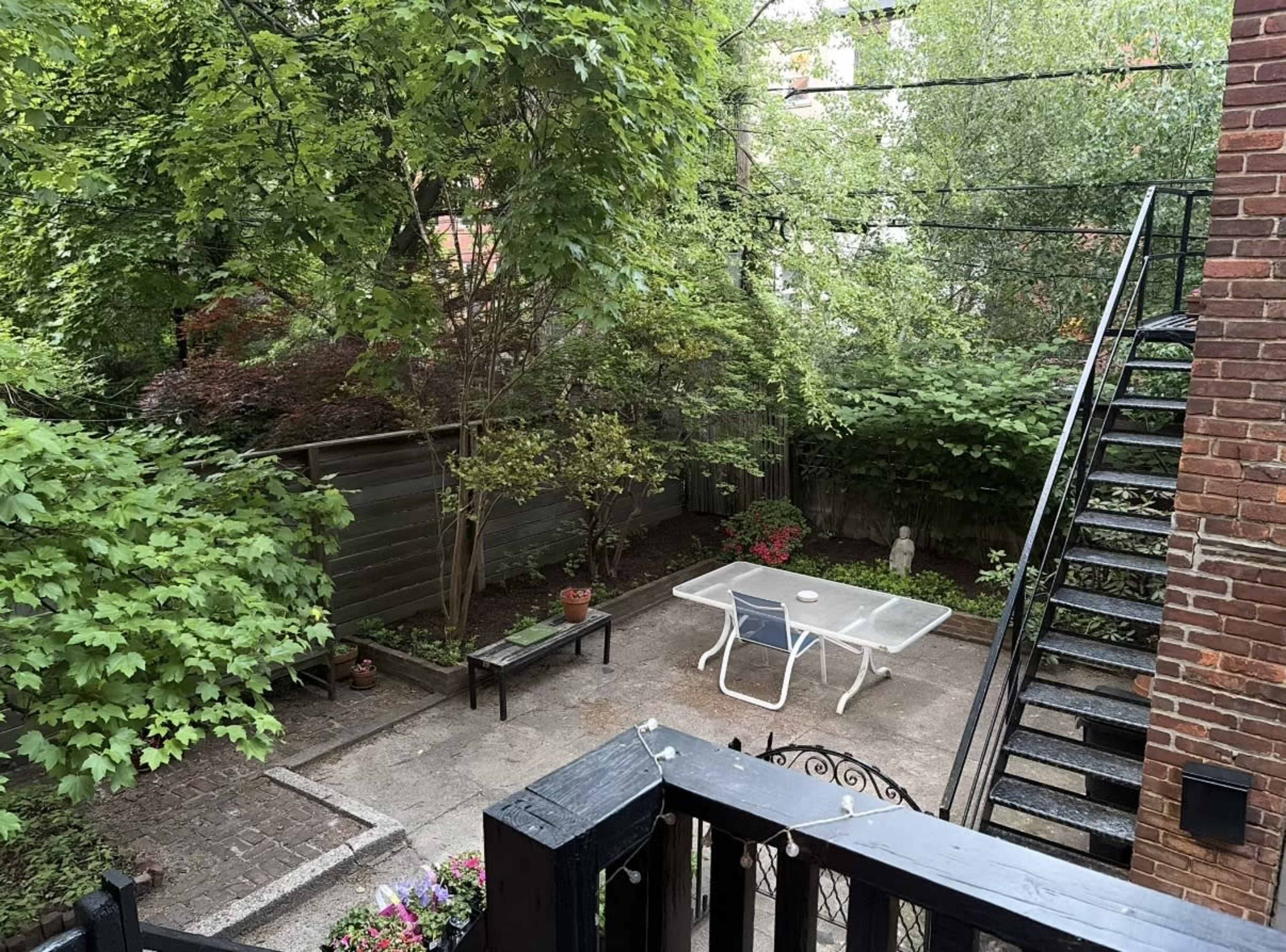 The image shows a small, green backyard with a patio table and chairs surrounded by trees and plants, along with a staircase leading to an upper level.