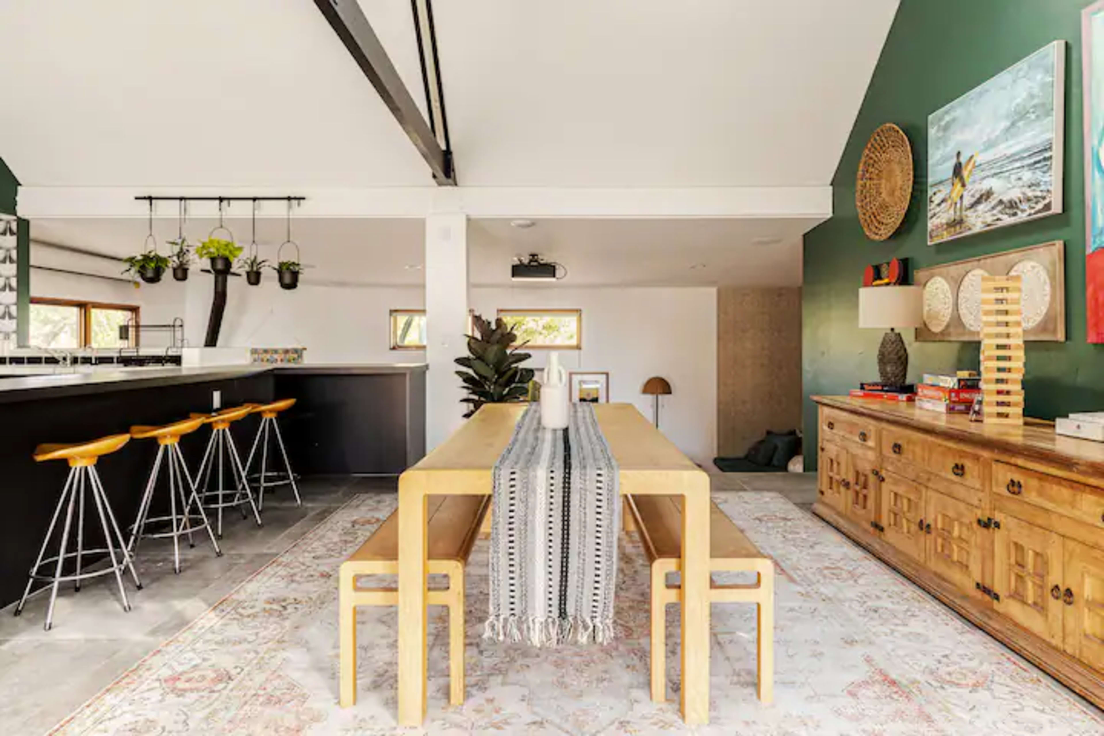The image shows a modern dining area featuring a long wooden table with benches, surrounded by green walls, and adjacent to a kitchen bar with tall stools.