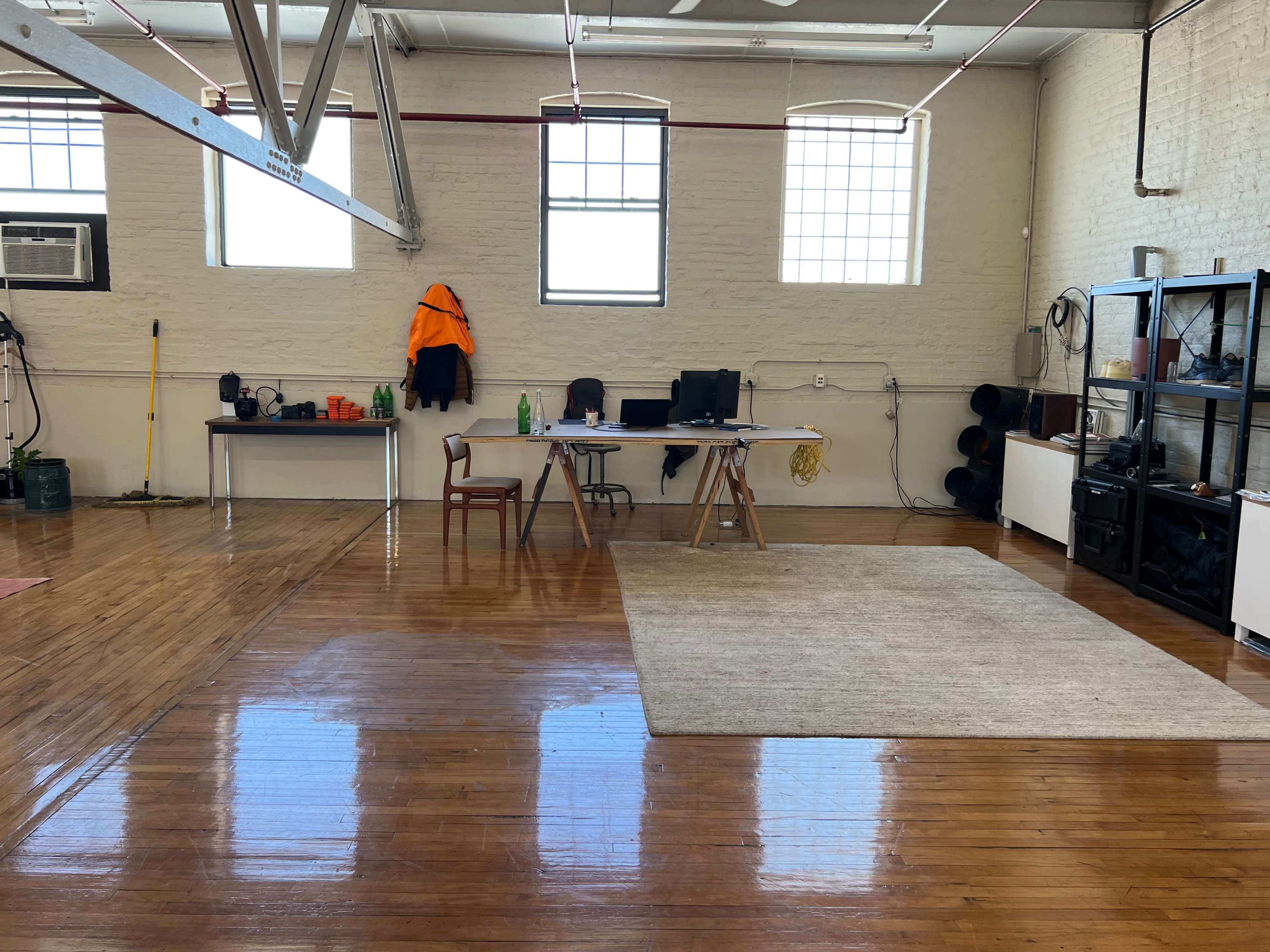 A spacious, well-lit room with polished wooden floors, featuring a desk with a computer, a chair, and various items on a table, along with a rug in the center.