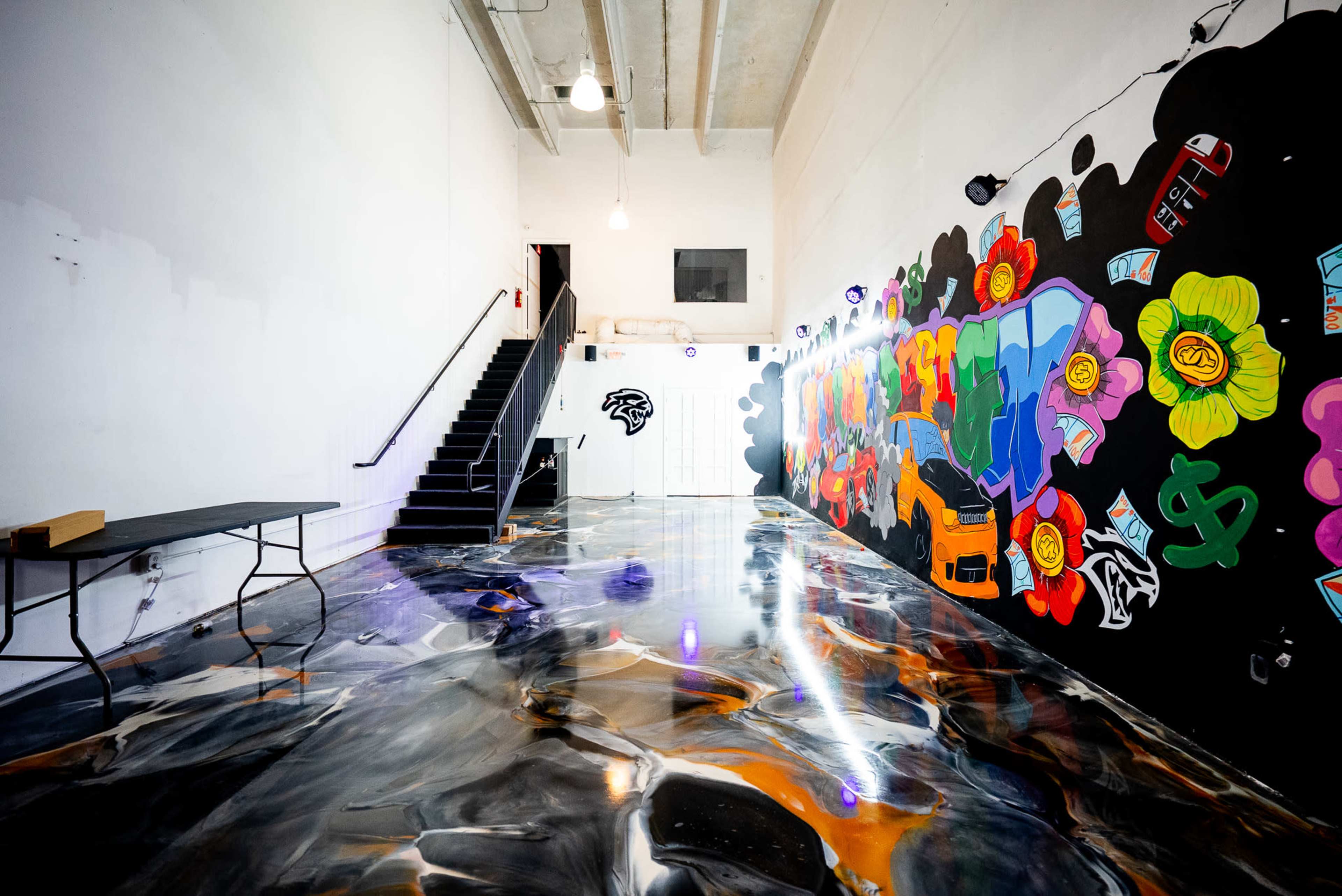 The image shows a contemporary indoor space featuring a glossy floor and a colorful mural on one wall, with a staircase in the background.