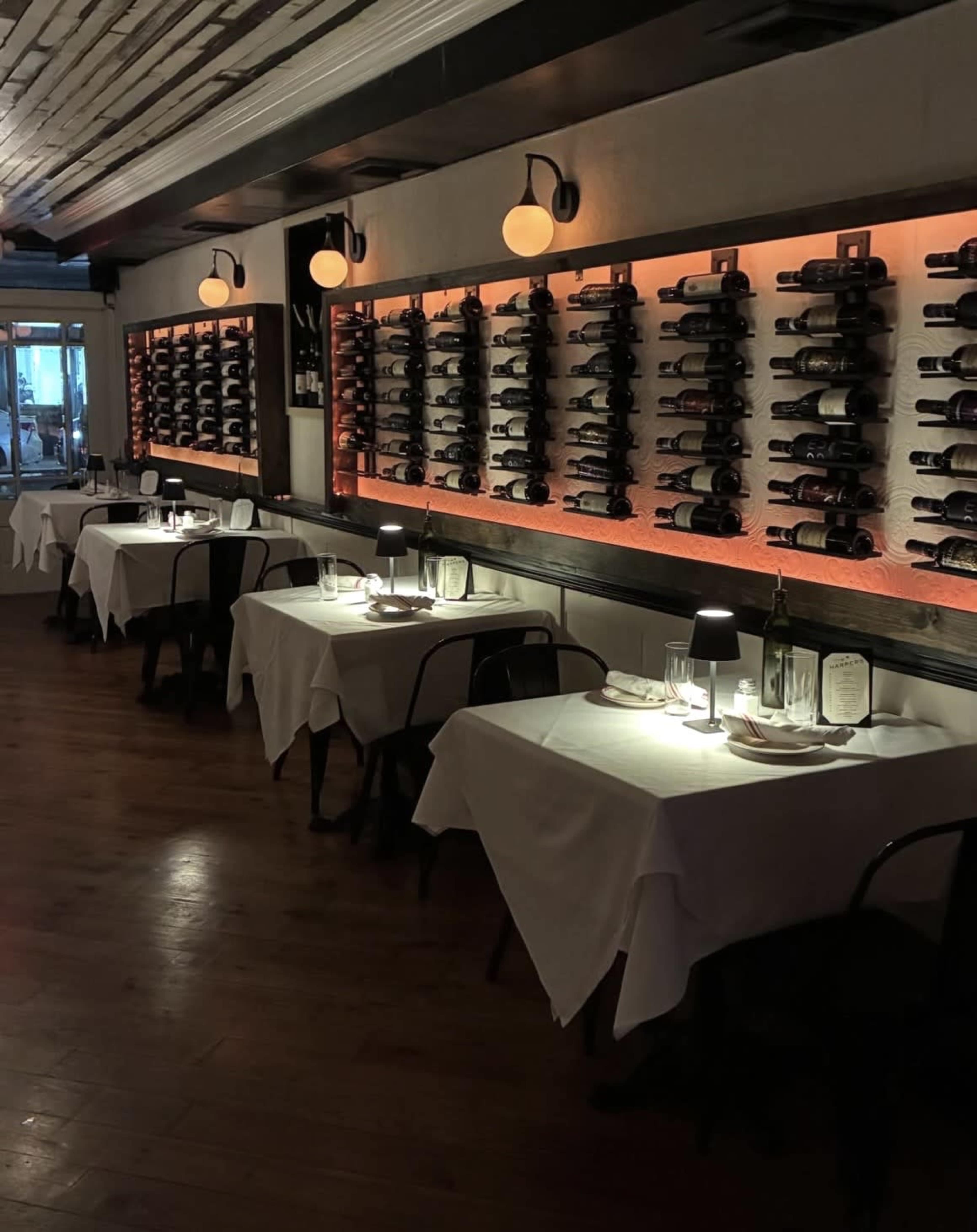 The image shows a dimly lit restaurant interior with white tablecloths, small lamps on each table, and a wall displaying an extensive wine collection.