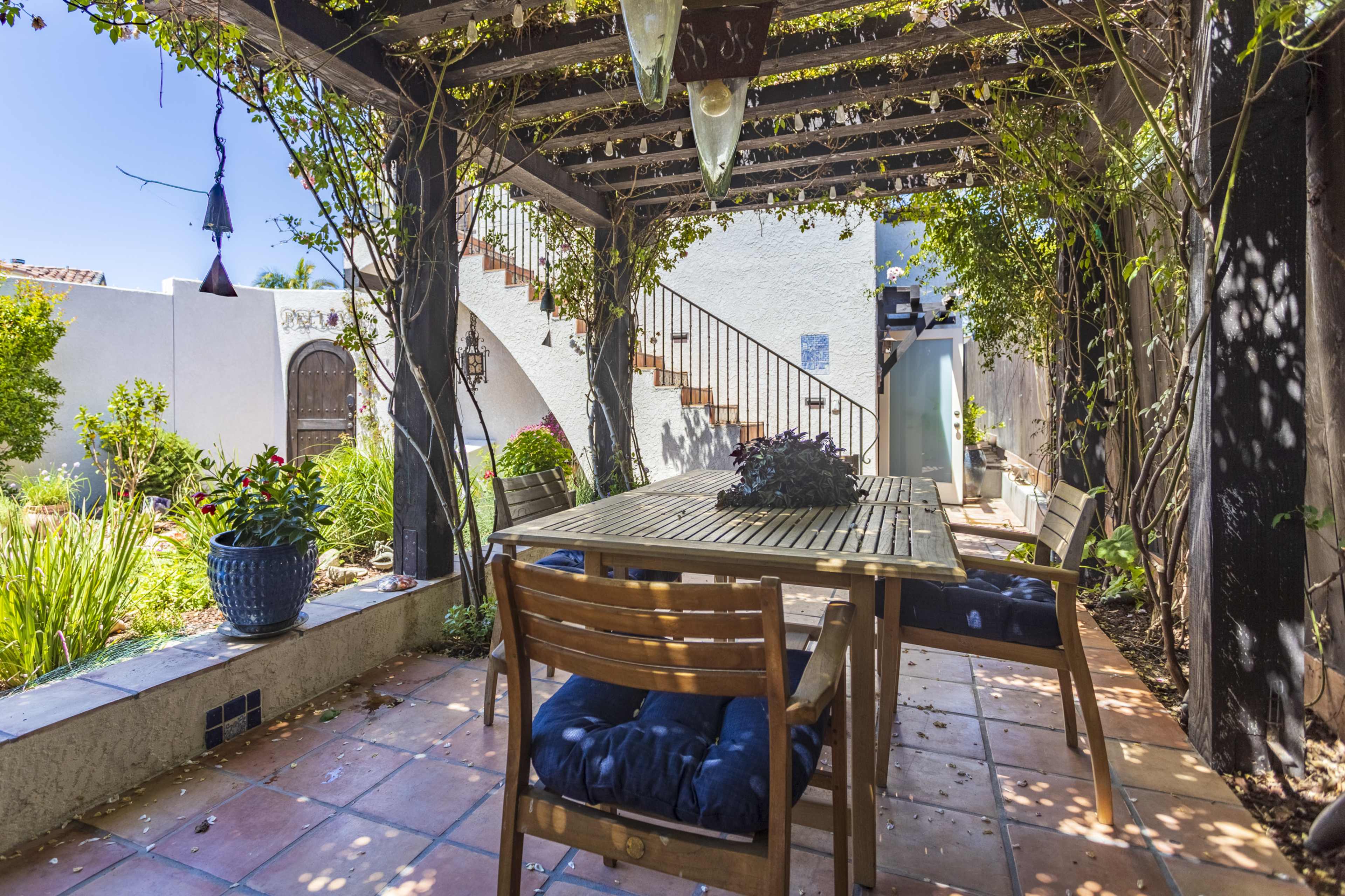 A wooden dining table with chairs is positioned under a pergola adorned with climbing plants, leading to a white-walled staircase in a garden setting.