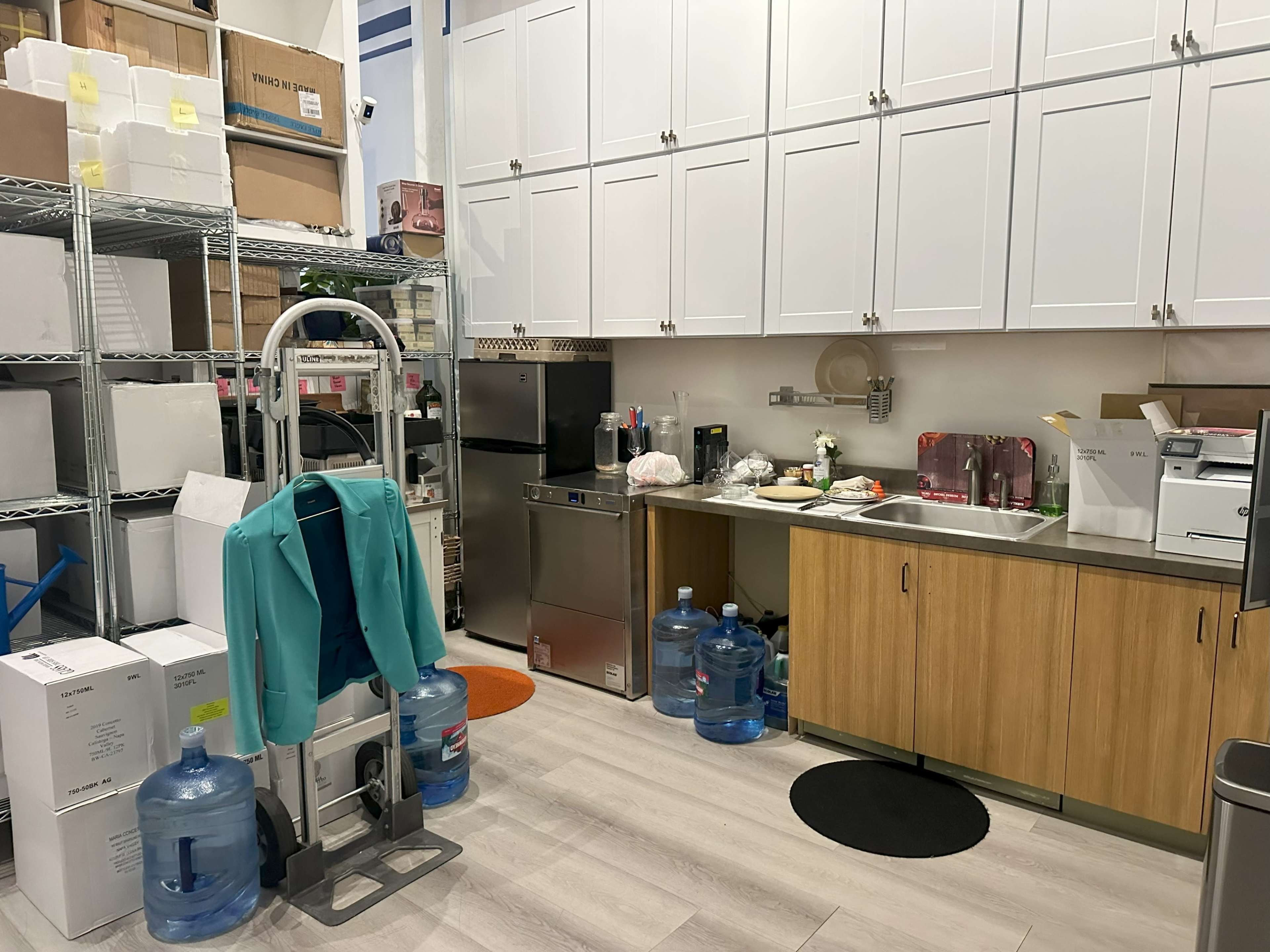 A small kitchen area with a sink, cabinets, a countertop filled with various items, and storage shelves holding boxes and water dispensers.