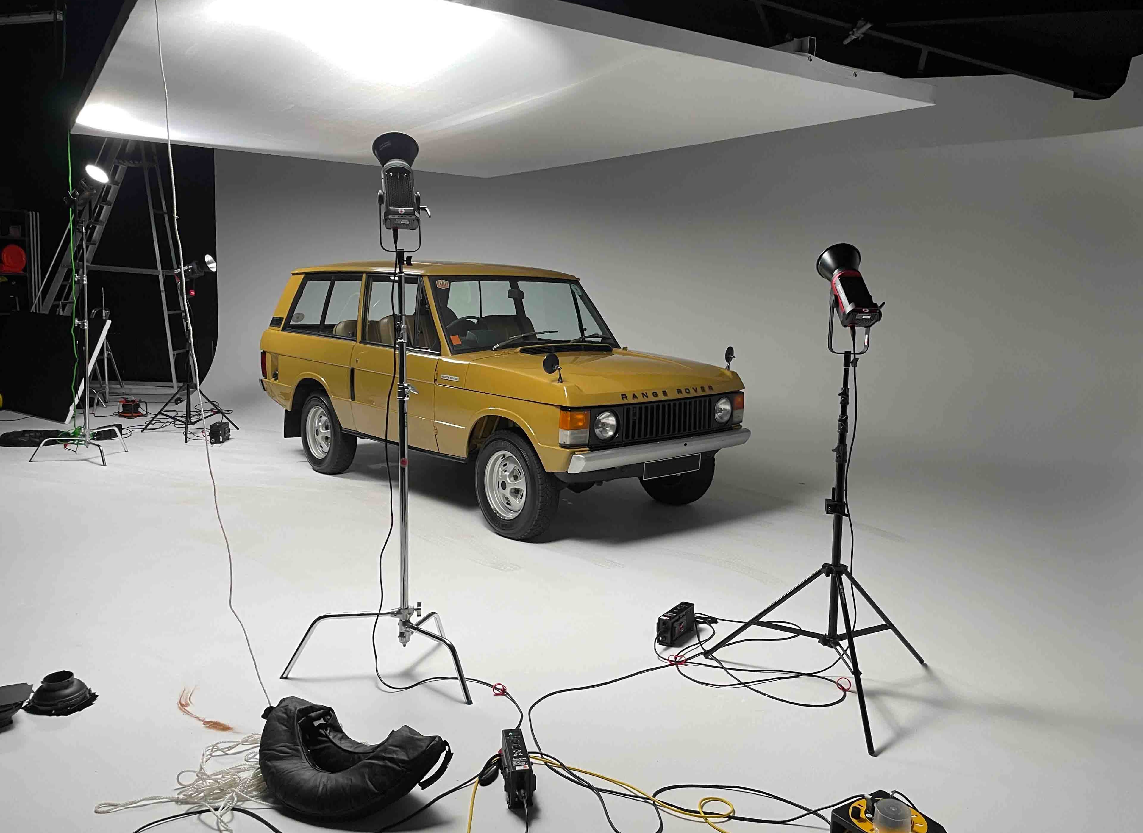 A vintage yellow SUV is positioned in a photography studio surrounded by lighting equipment and a backdrop.