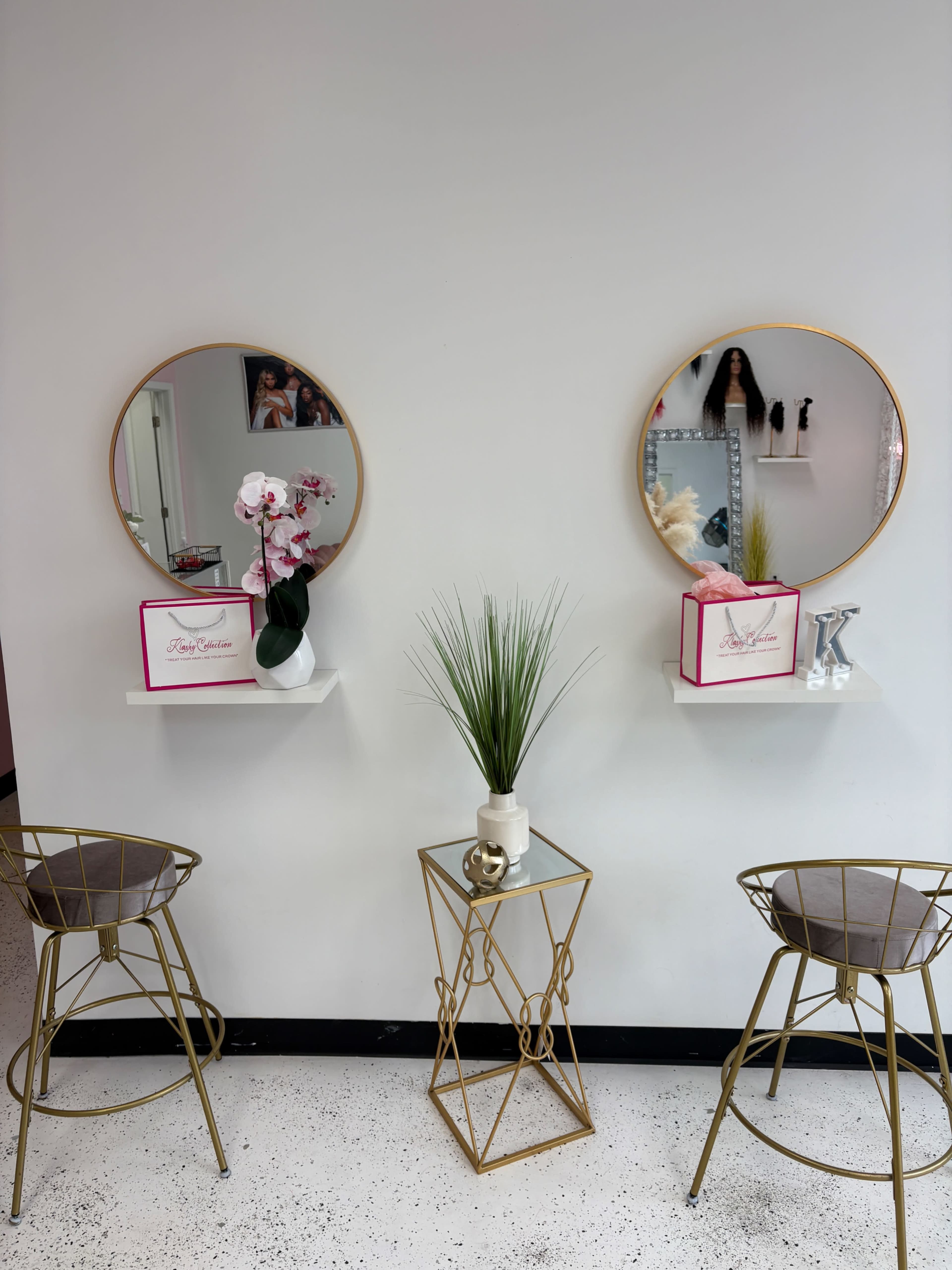 The image shows a minimalist salon space with two round mirrors mounted on the wall above small white shelves, flanked by a decorative plant and a gold side table with two stylish bar stools.