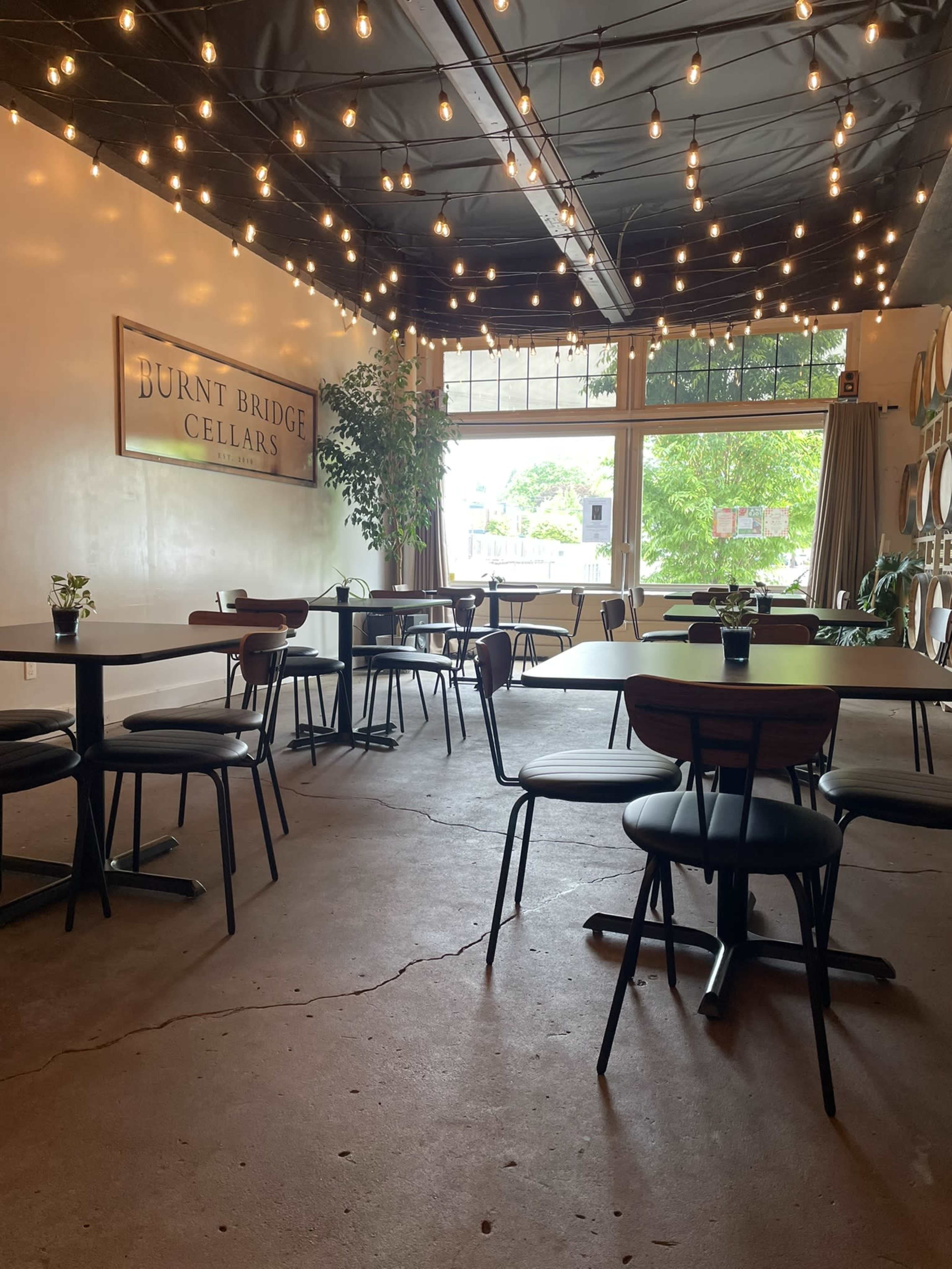 The image shows a spacious interior of a restaurant or tasting room with wooden tables, chairs, string lights overhead, and large windows letting in natural light.