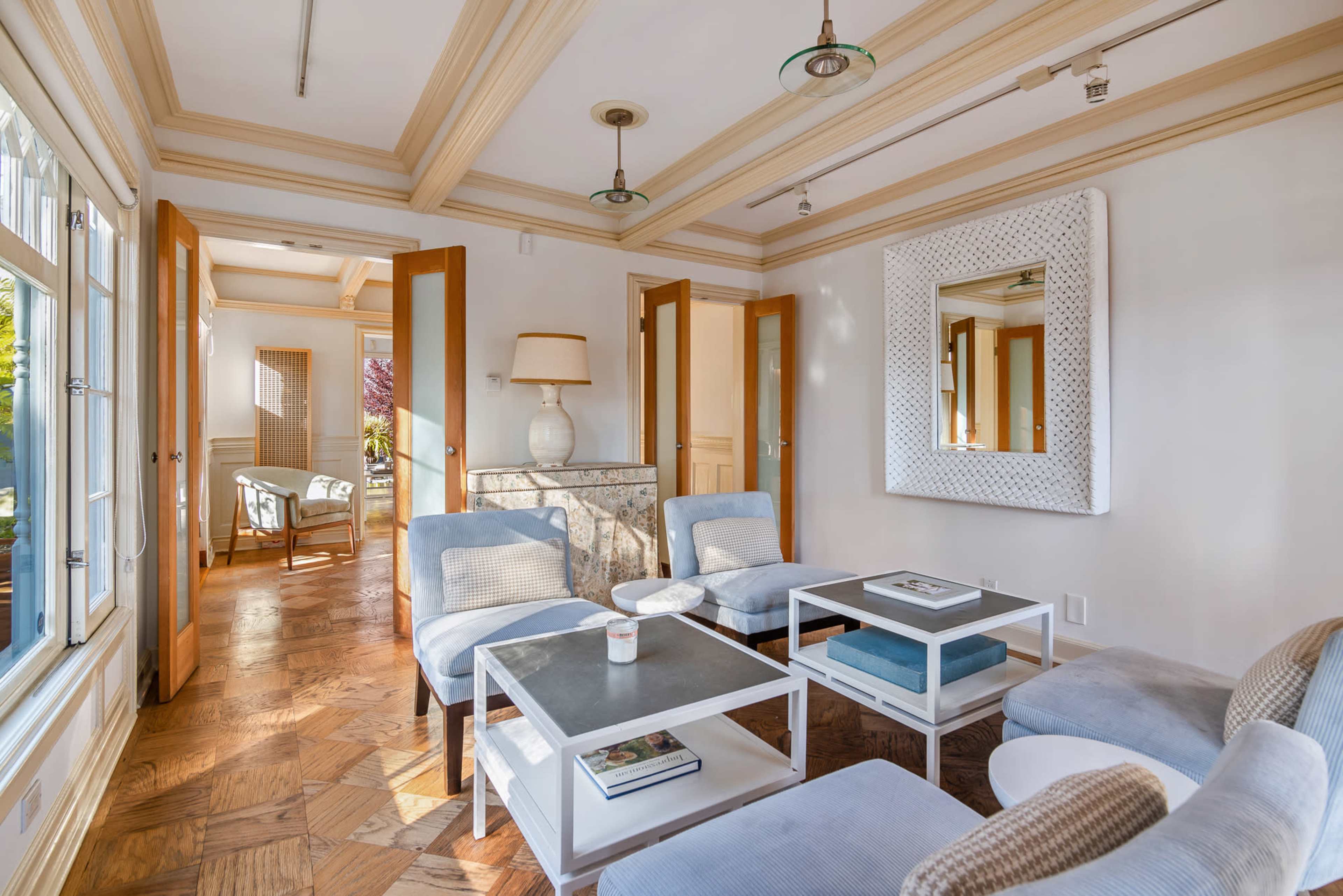The image depicts a living room with a light-colored seating area, a coffee table, and a large mirror on the wall, framed by wooden doorways leading to other spaces.