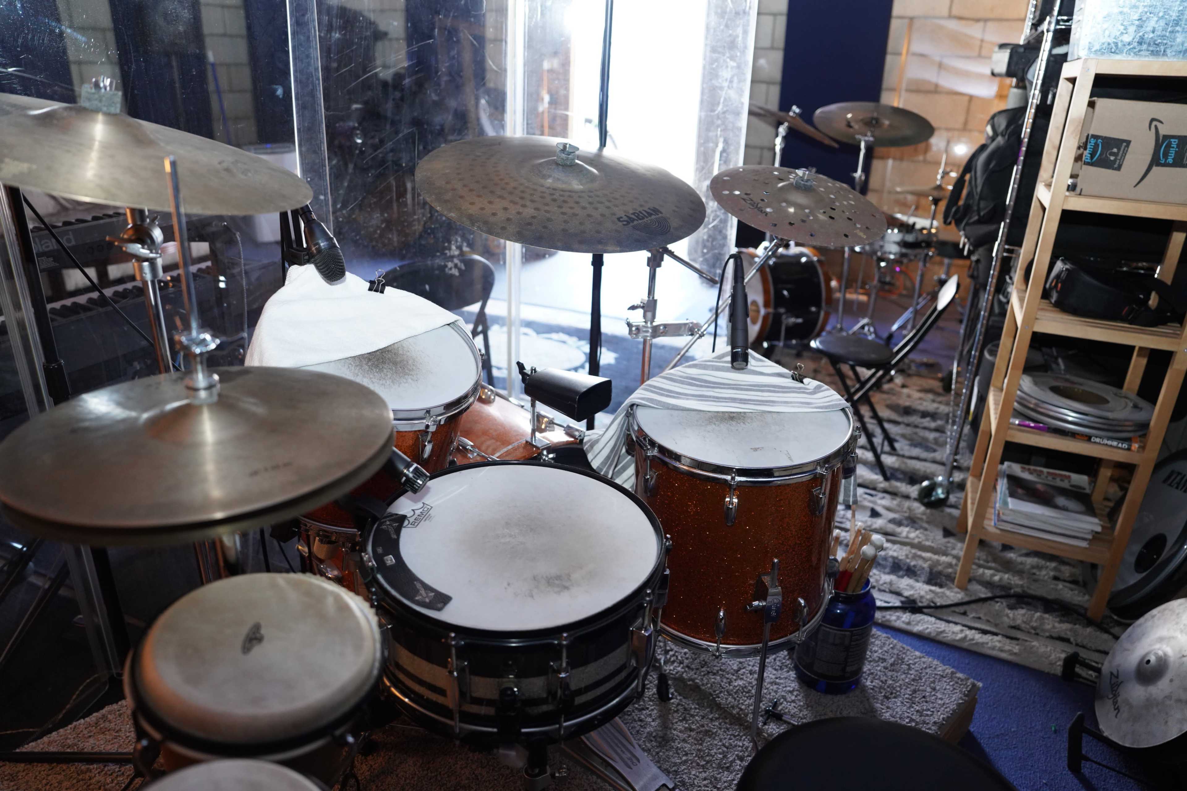 The image shows a set of drums in a recording studio, featuring a snare drum, toms, cymbals, and various musical equipment in the background.