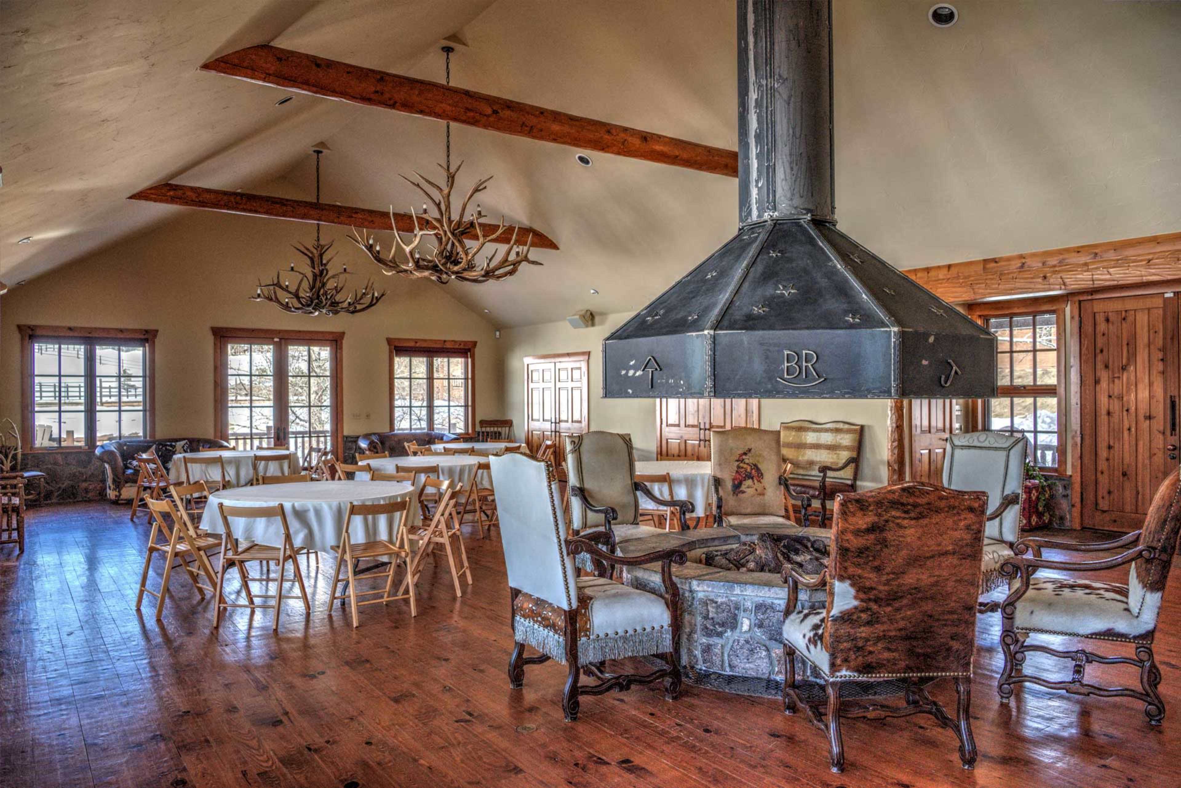 The image shows a spacious communal area featuring wooden flooring, several round tables with chairs, and a central stone fireplace surrounded by a seating arrangement.